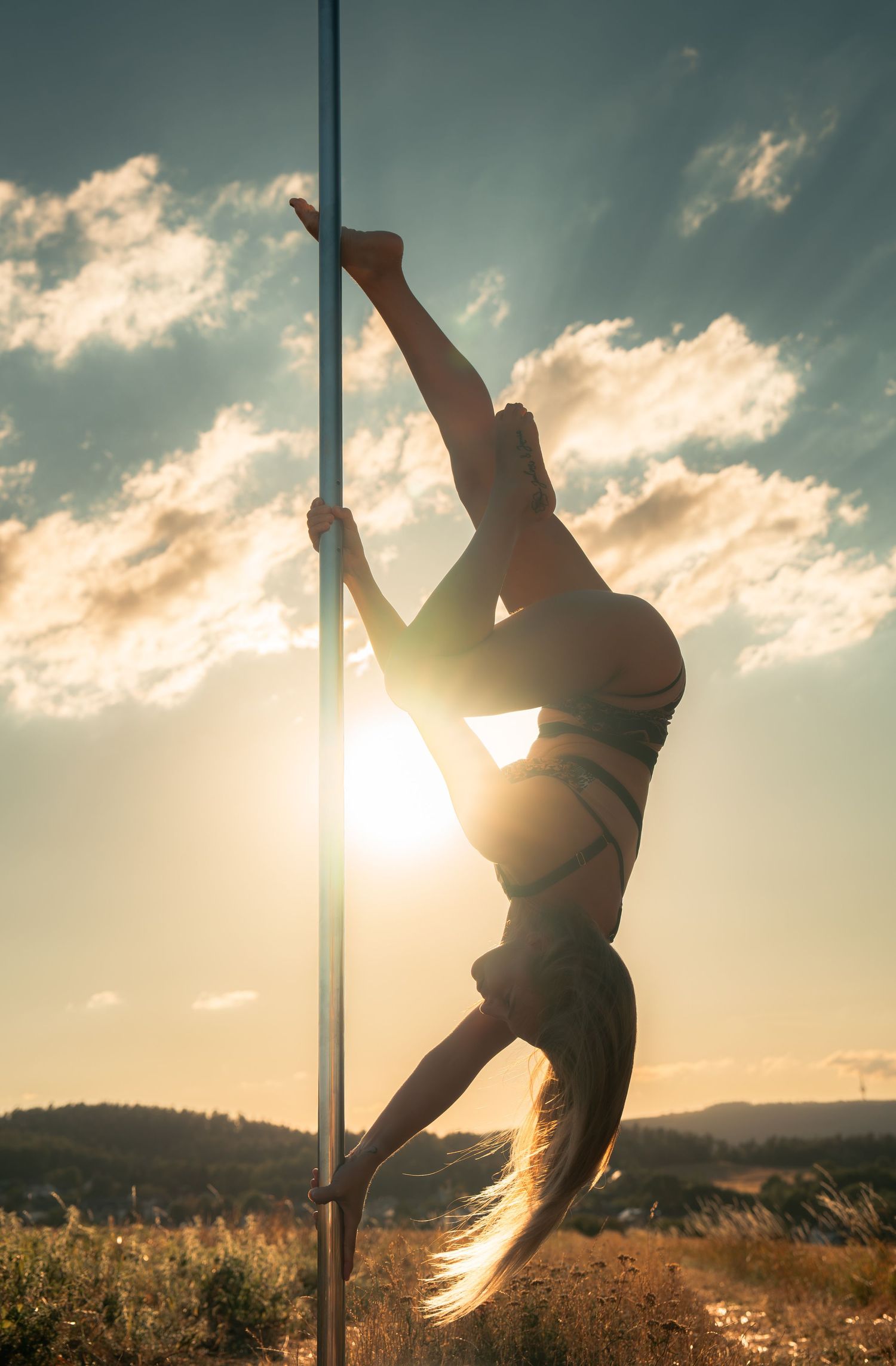 Silhouette of a woman performing pole dance outdoors at golden sunset with dramatic sky and sun rays.