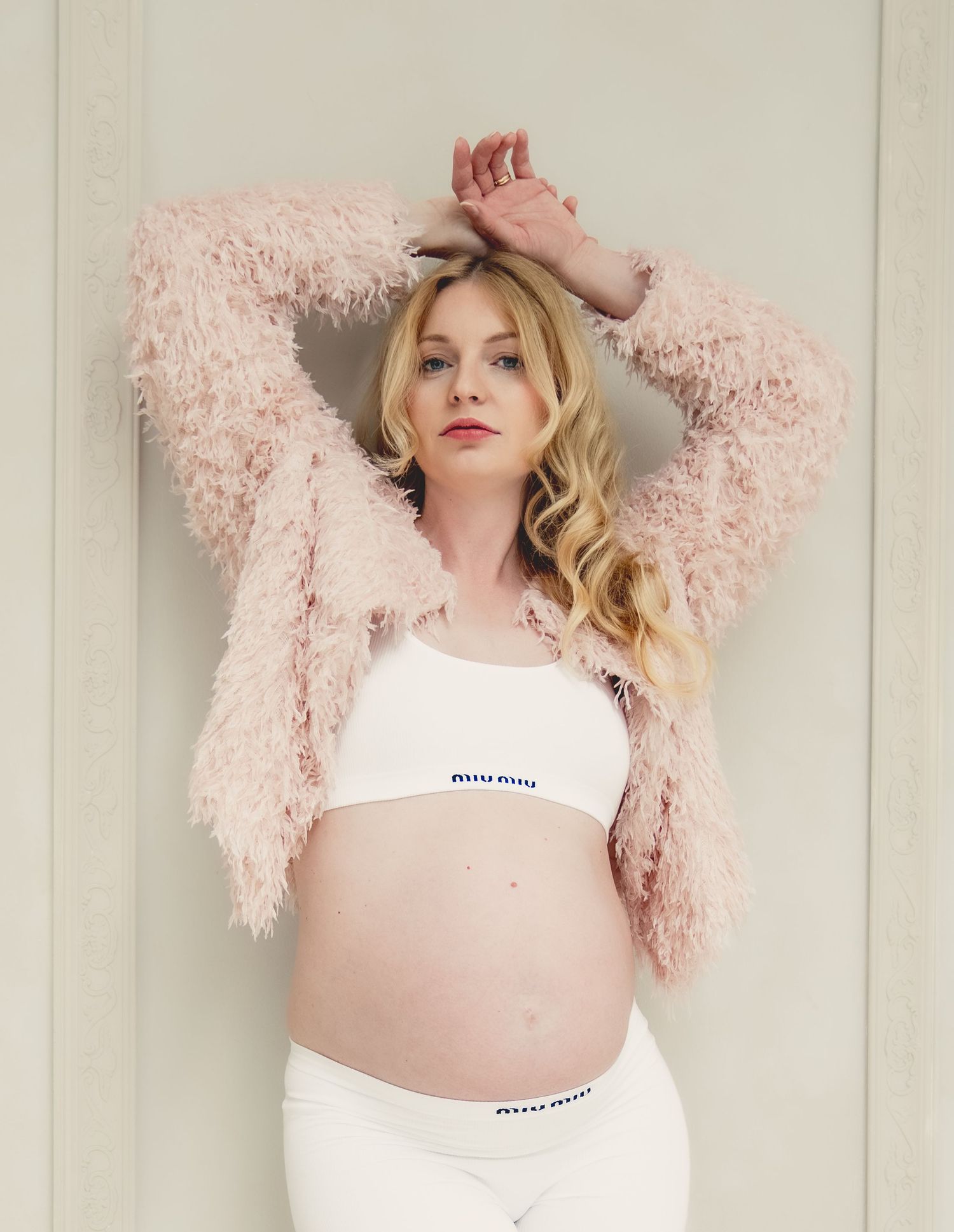 Pregnant woman in white Miu Miu bralette and underwear with pink fluffy jacket, posing with arms raised.