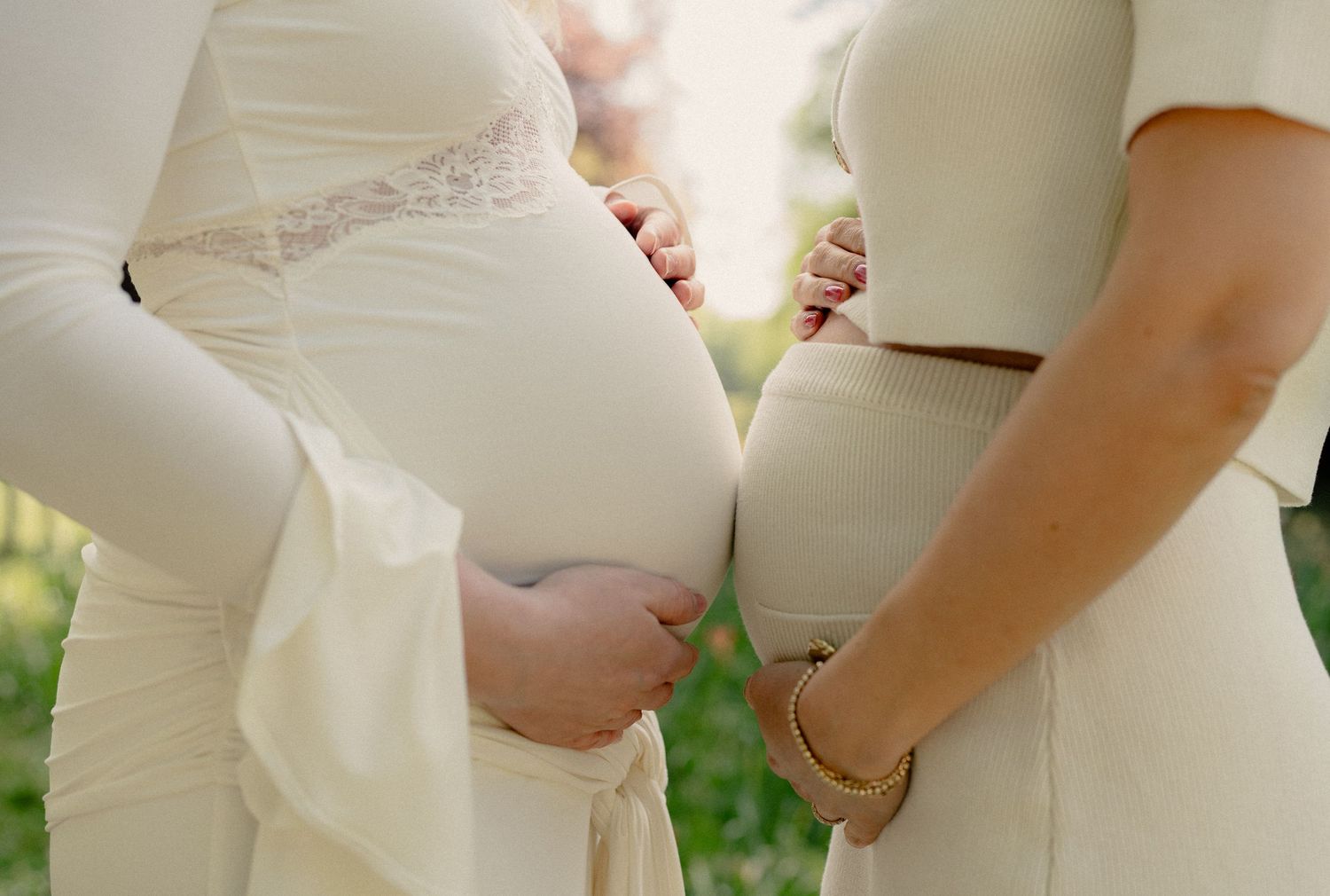 Two pregnant women in white outfits standing close together, both cradling their baby bumps outdoors.