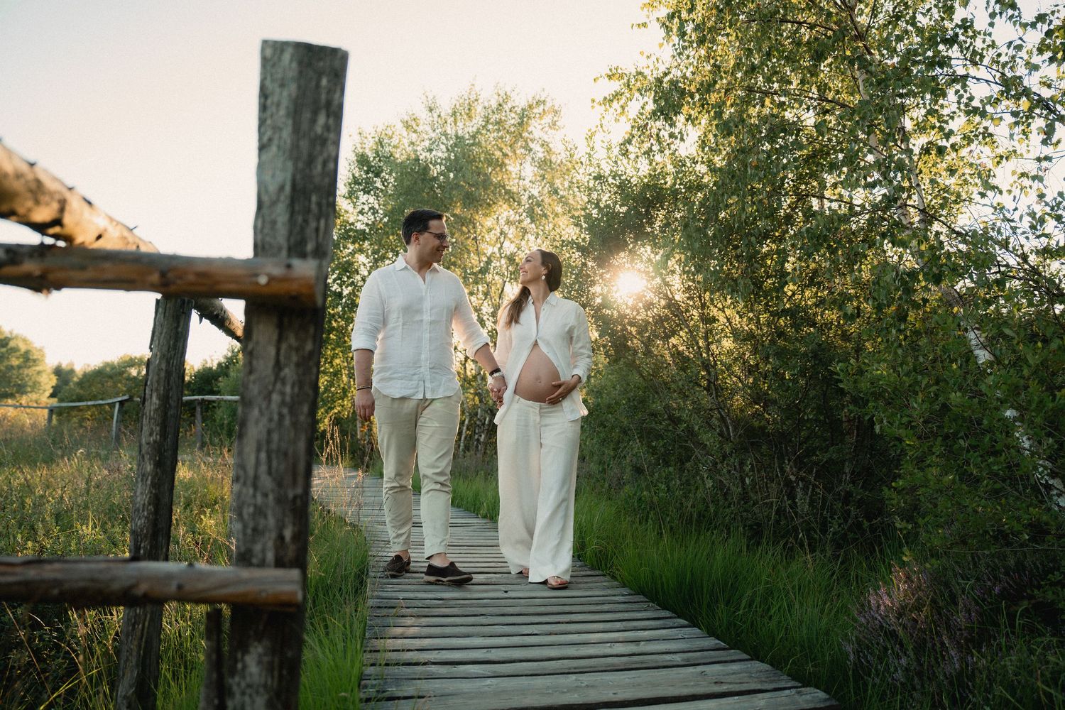 Pregnant couple holding hands on a wooden bridge surrounded by lush greenery in golden sunlight.