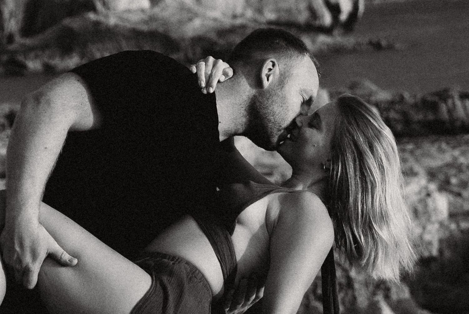 Black and white photo of a romantic couple sharing a passionate kiss outdoors near rocky terrain.