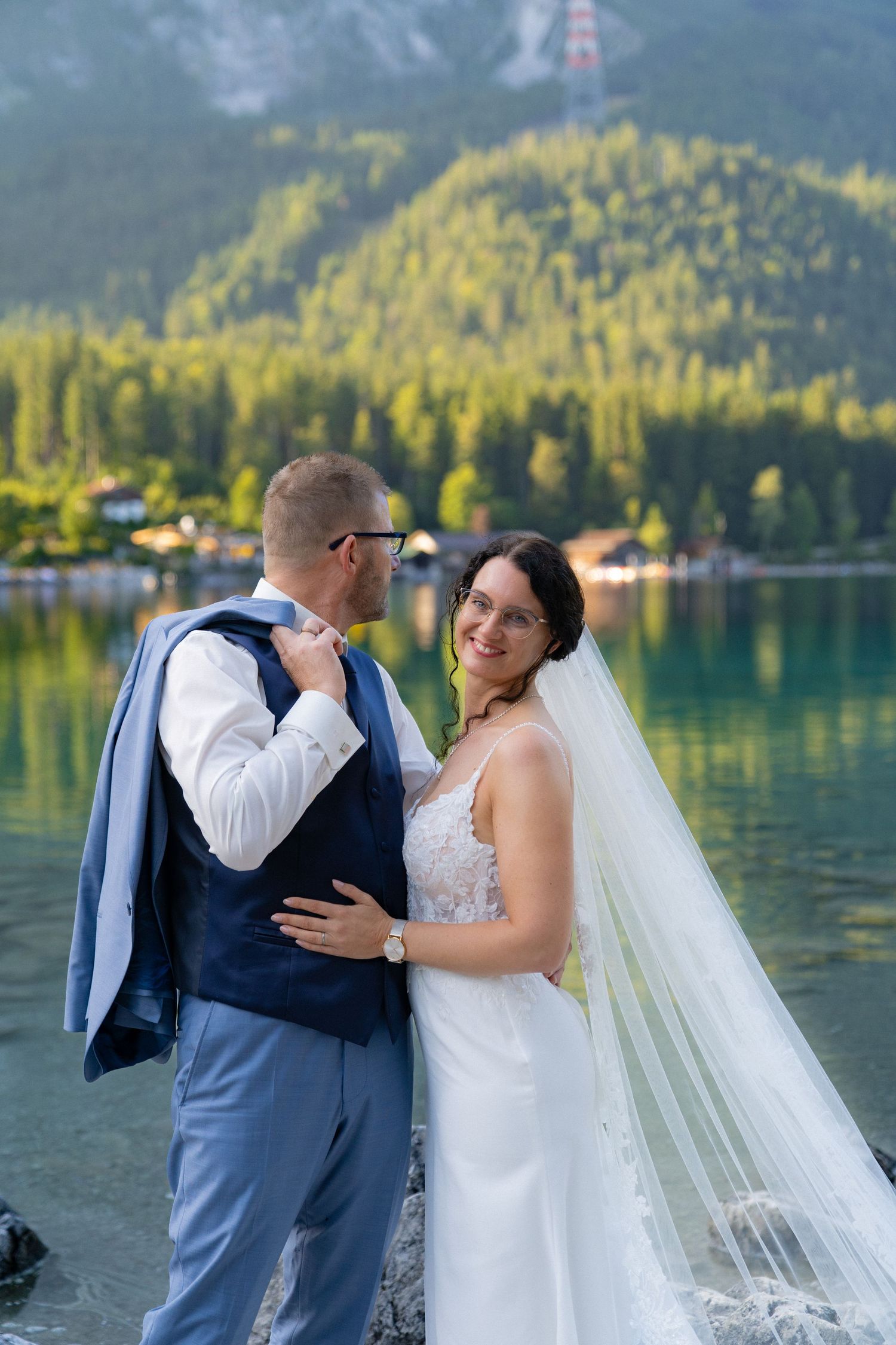 A couple in wedding attire shares romantic moments by a scenic mountain lake with evergreen trees in the background.