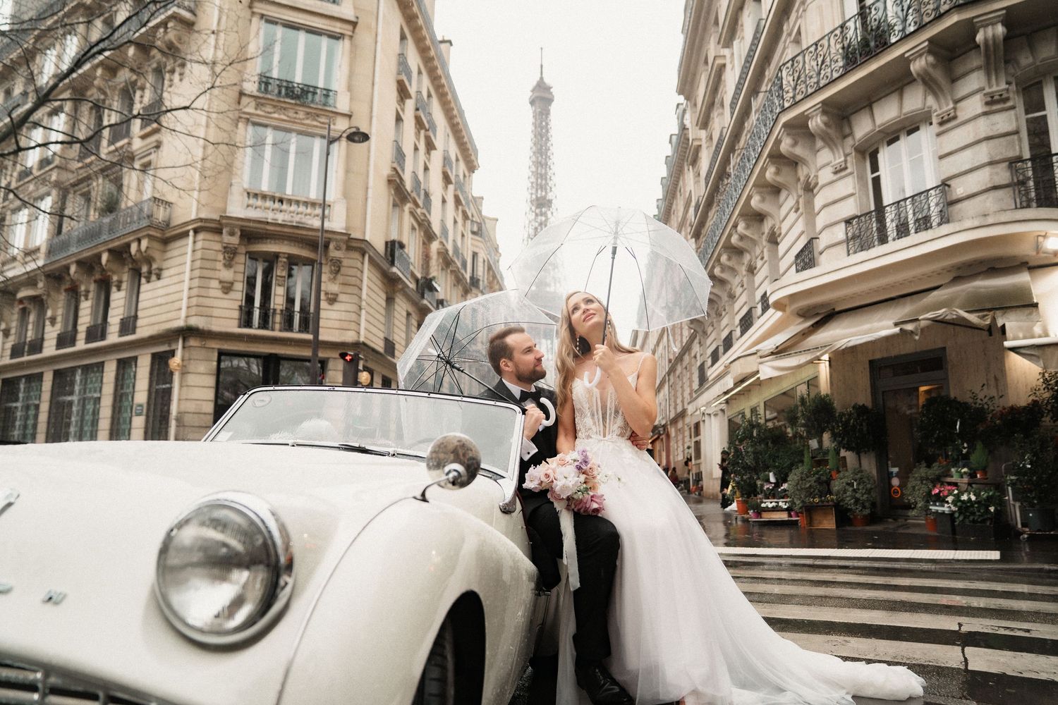 A romantic wedding couple poses near the Eiffel Tower in Paris with vintage cars and elegant architecture in view.