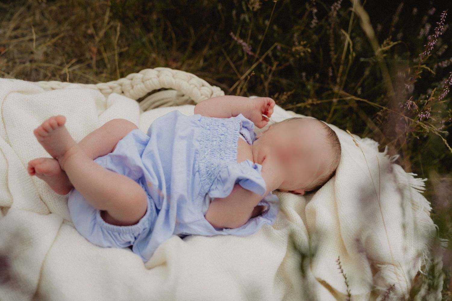 Peaceful newborn baby sleeping outdoors on white blanket in blue romper surrounded by natural grass.