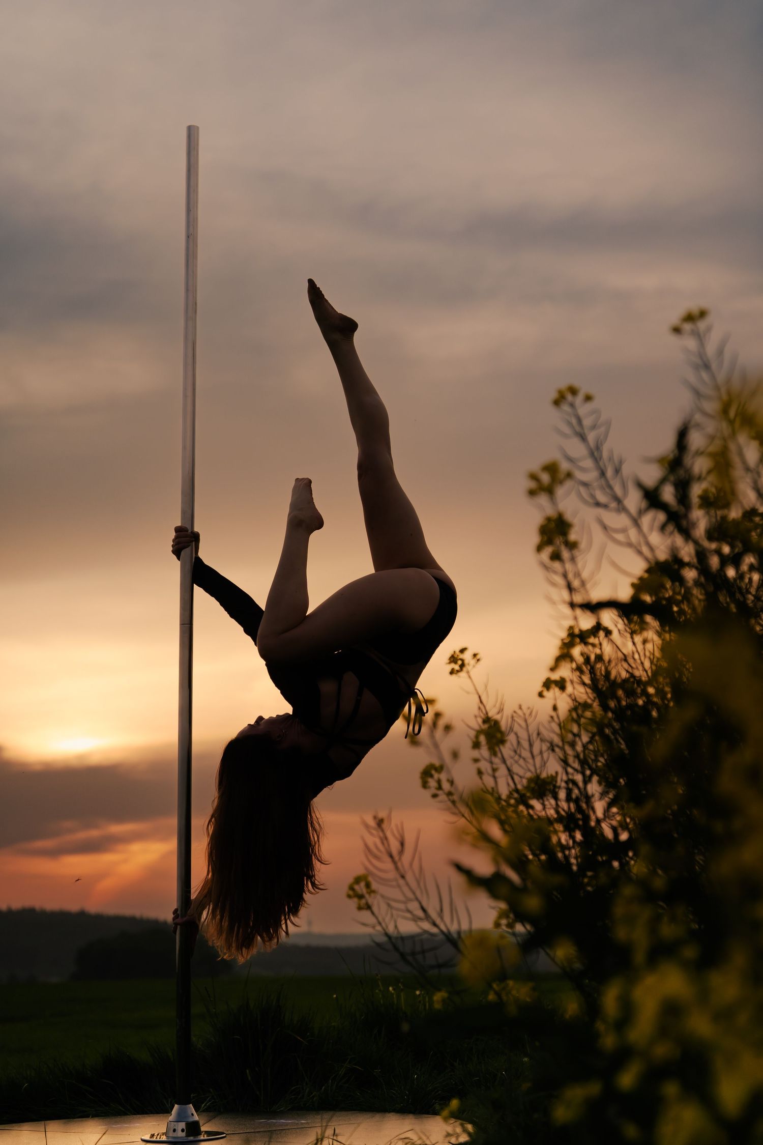 Silhouette of a woman performing pole dance outdoors against a dramatic sunset sky with golden hues.