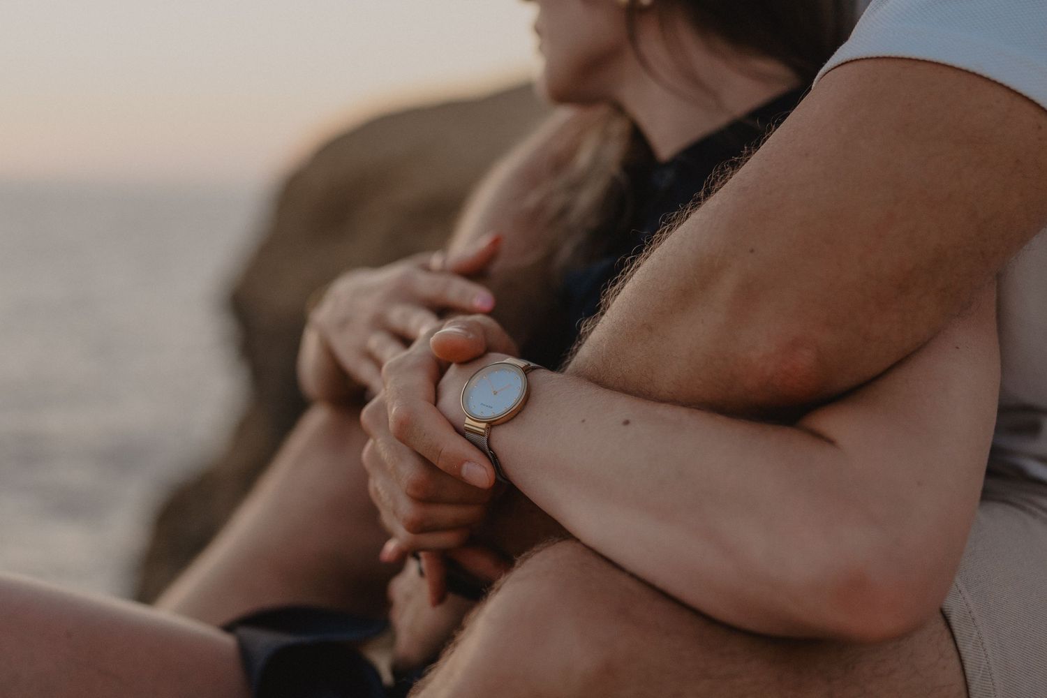 Couple embracing tenderly at sunset by the ocean, holding hands with a white watch visible on wrist.