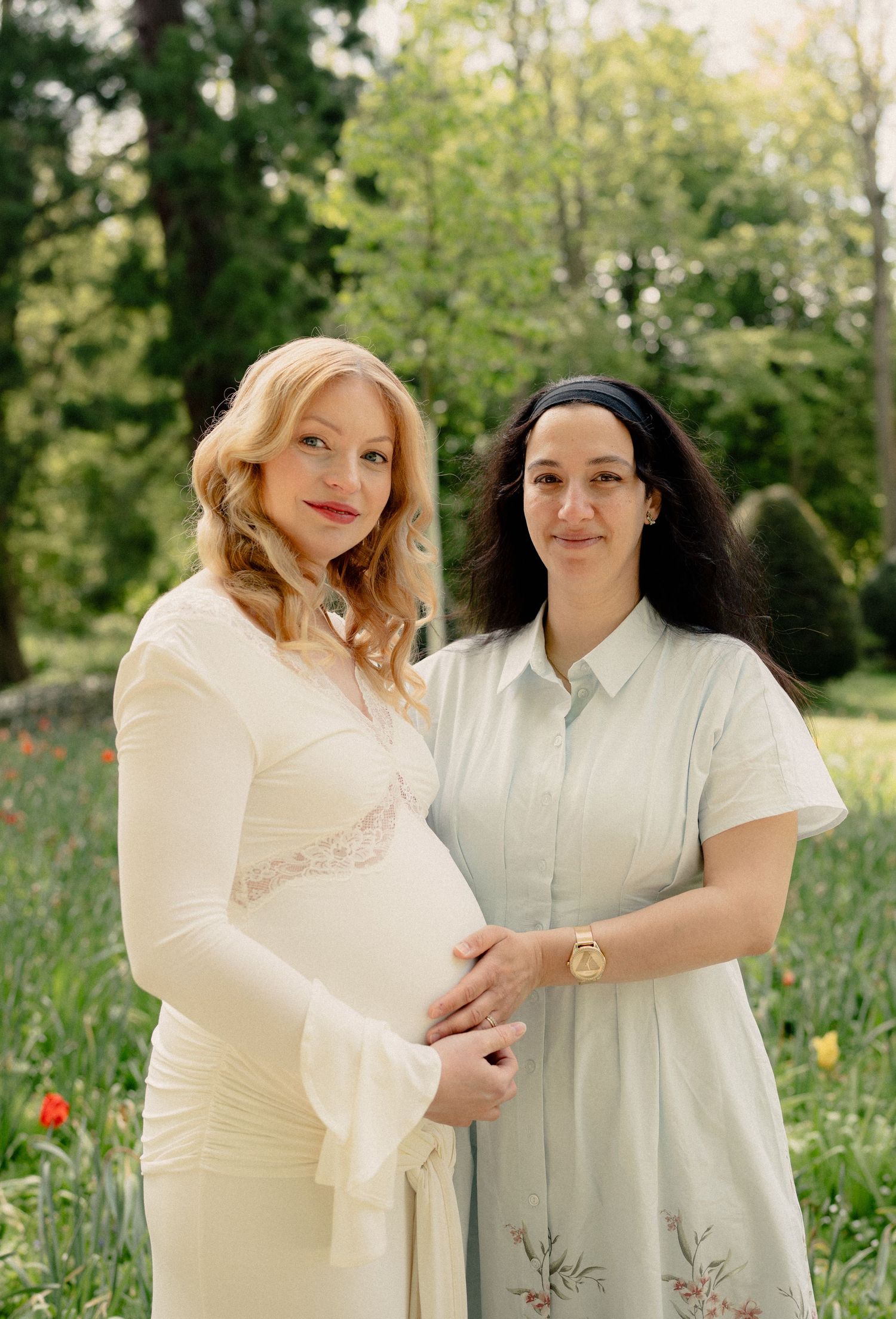 Two women pose in a lush garden, one in a white maternity dress cradling her baby bump, surrounded by colorful tulips.
