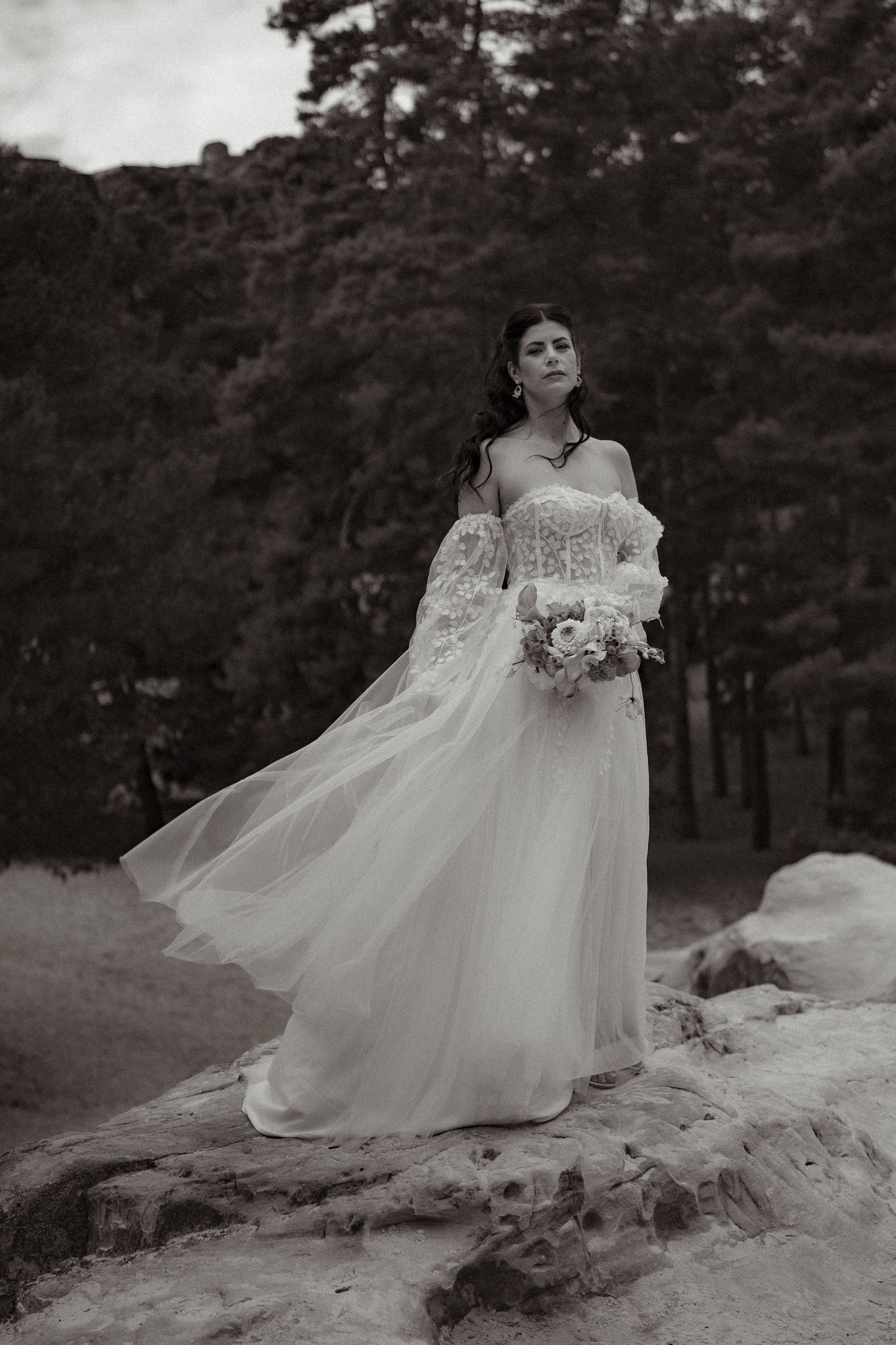 A bride in a flowing white gown poses gracefully on rocky terrain surrounded by pine trees.