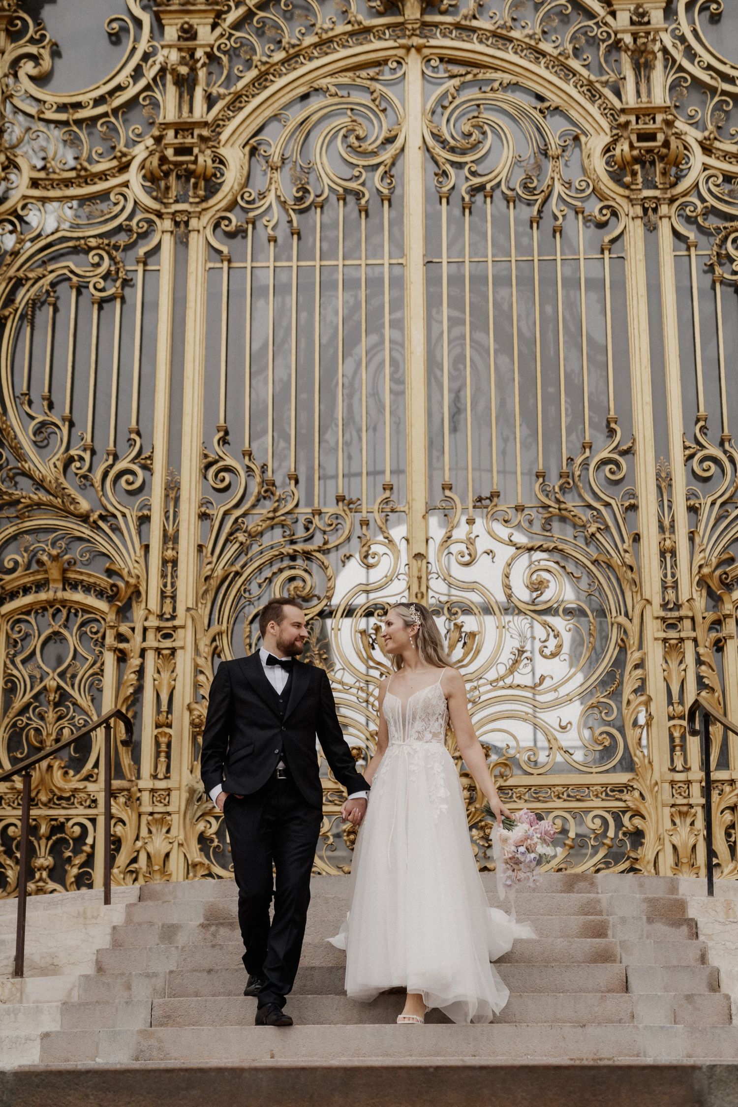 Couple descending ornate golden baroque staircase with elaborate metalwork and architectural details in a grand palace setting.