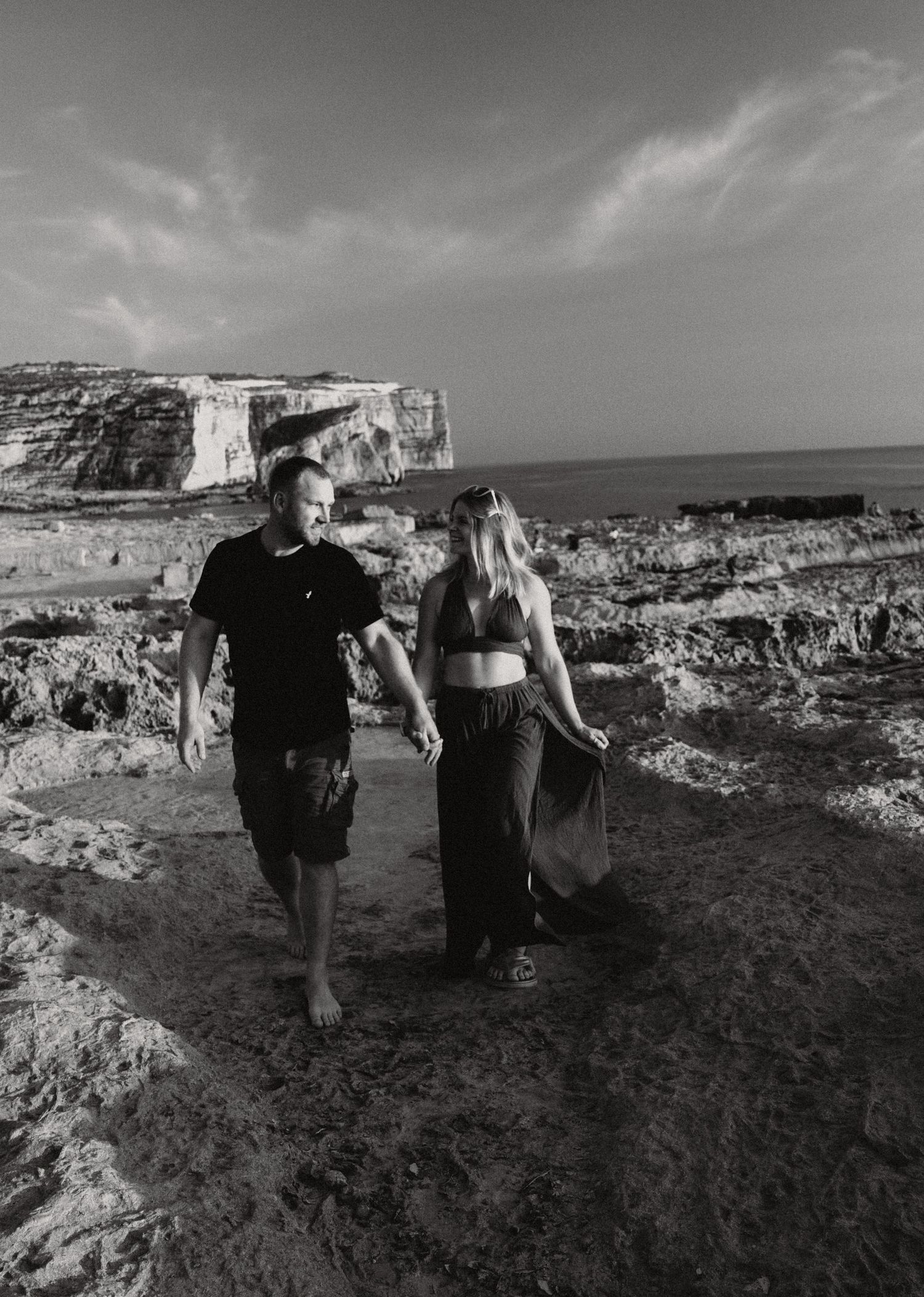 Black and white photo of a couple holding hands on rocky coastal cliffs with ocean and white cliffs in background.