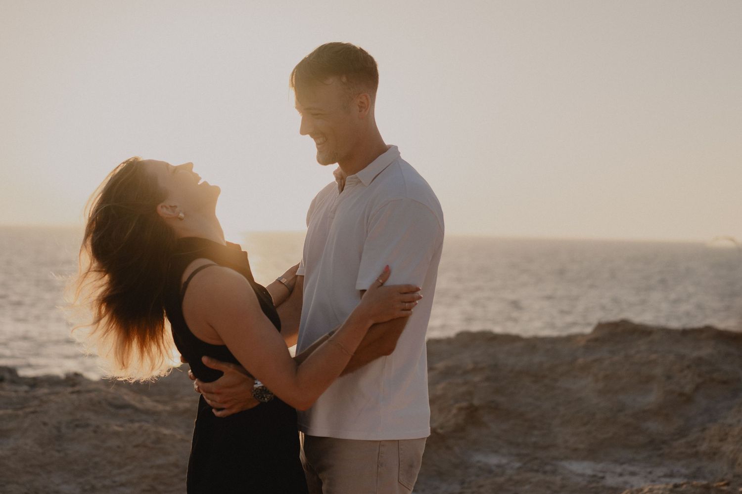 Couple embracing and smiling at sunset by the ocean on rocky coastal cliffs in a romantic golden hour moment.