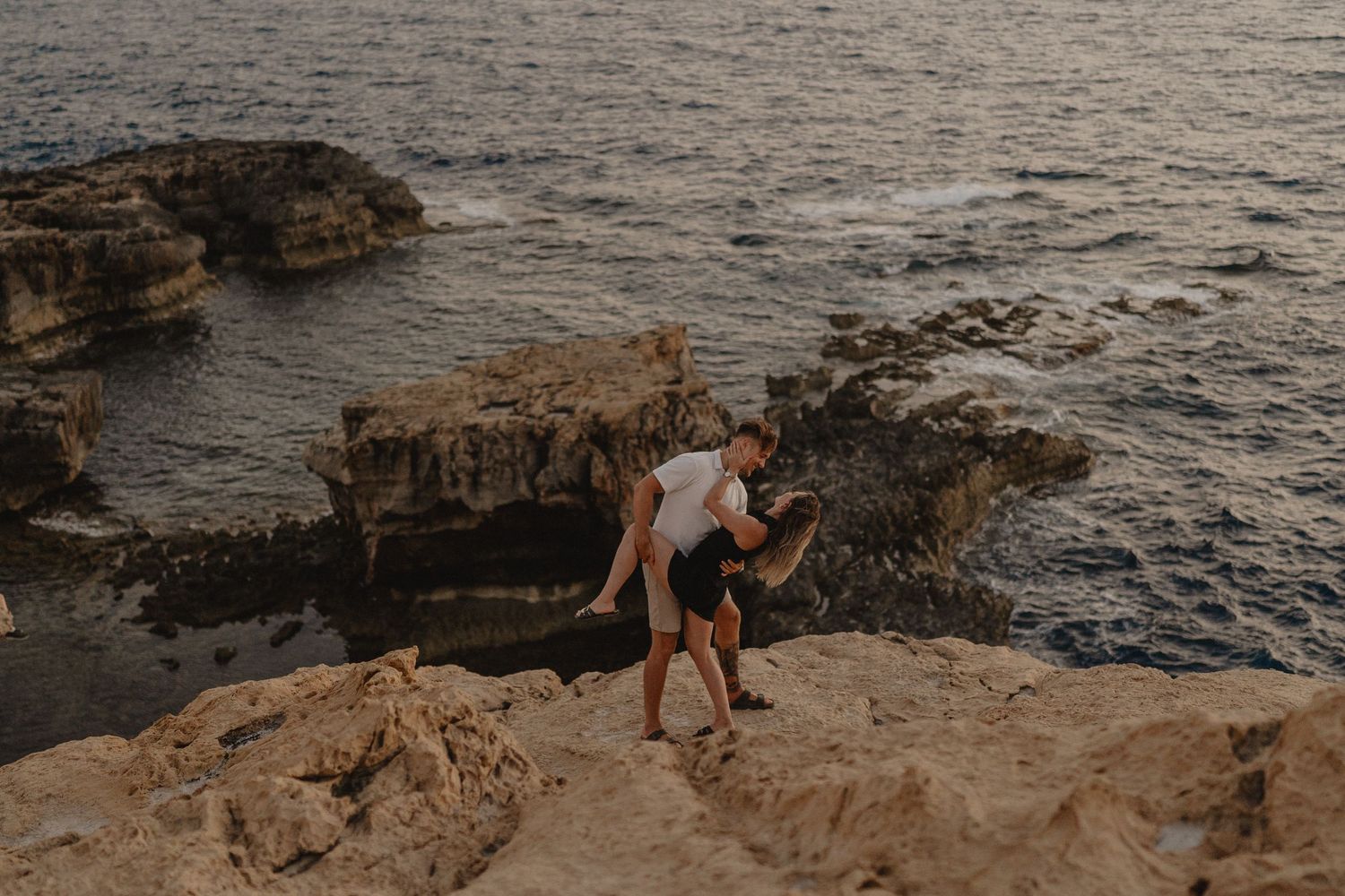 Couple embracing on rocky coastal cliffs at sunset with turquoise ocean waves in the background.