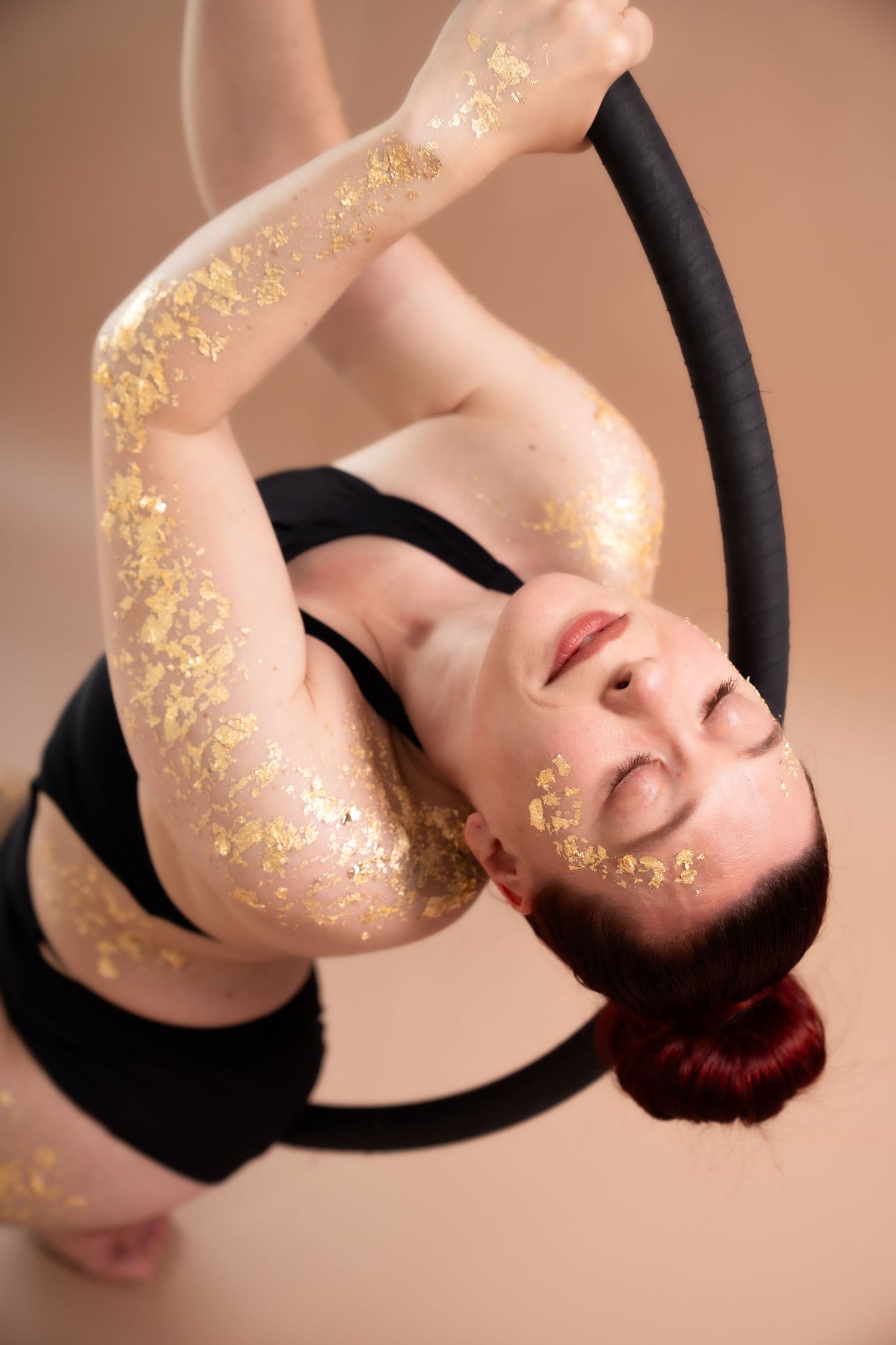 Aerial hoop performer with gold leaf body art poses gracefully in black athletic wear against beige background.
