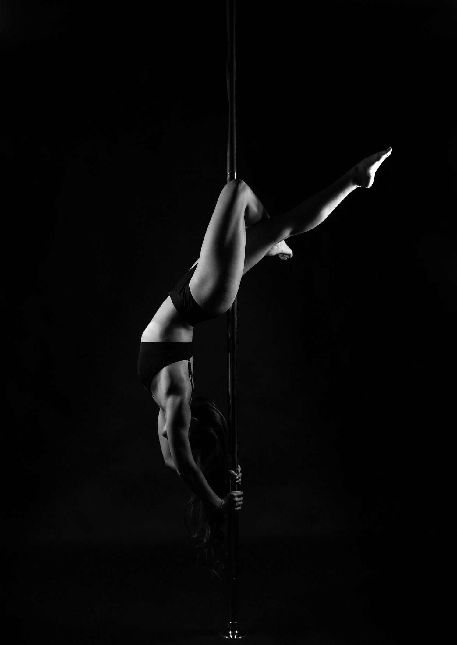 Black and white photo of a pole dancer performing an inverted split pose on a vertical pole against dark background.