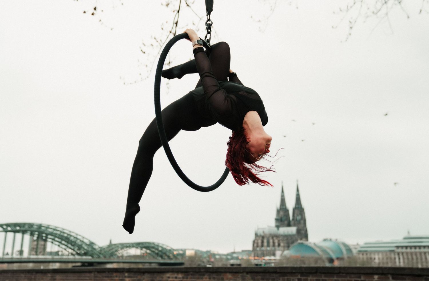Aerial hoop performer in black bodysuit hanging upside down outdoors with Cologne Cathedral in background.
