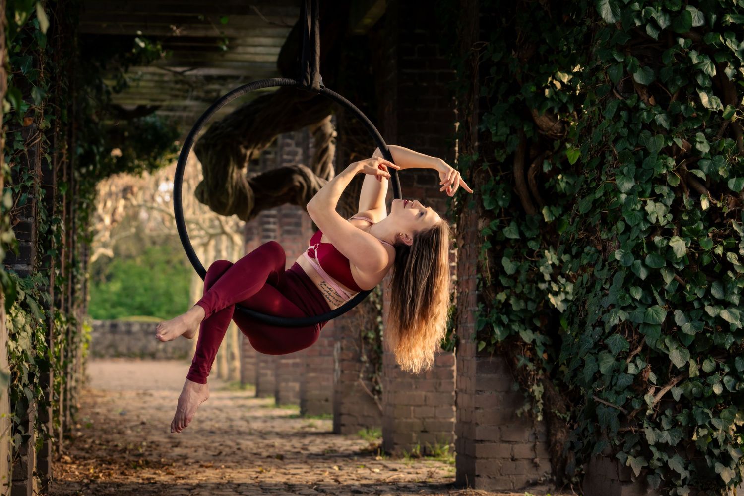 Woman in red performing aerial hoop dance outdoors under ivy-covered archway.