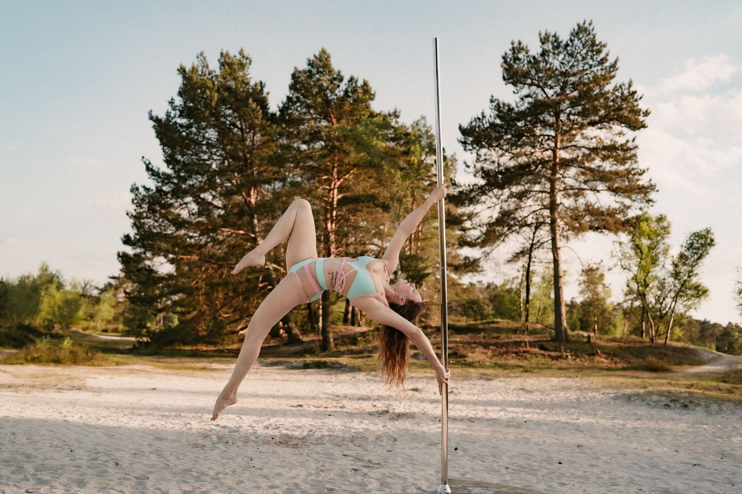 Woman performing pole dance outdoors on sandy beach surrounded by tall pine trees in summer daylight.