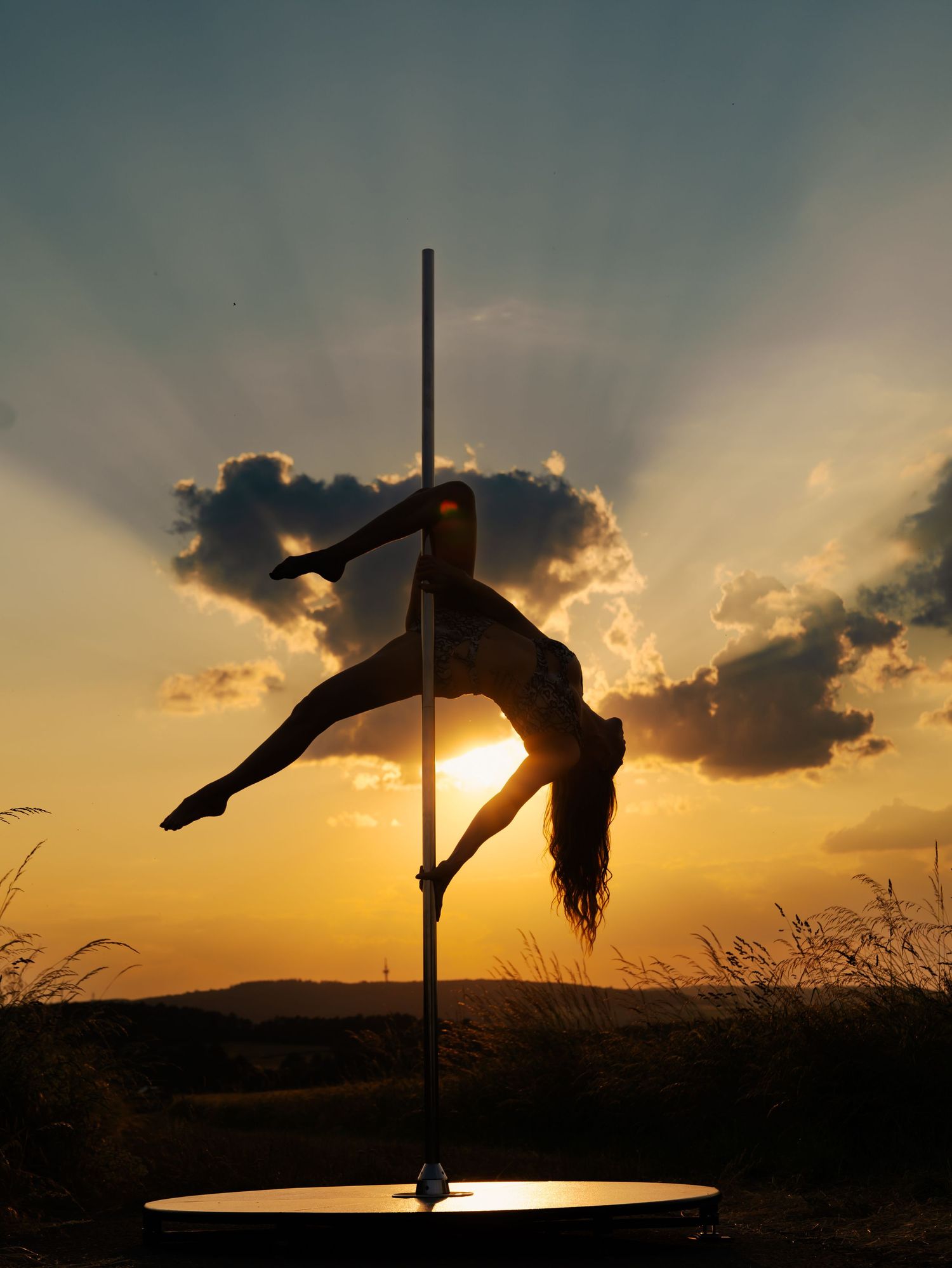 Silhouette of a woman performing pole dance against a dramatic golden sunset sky with glowing clouds.