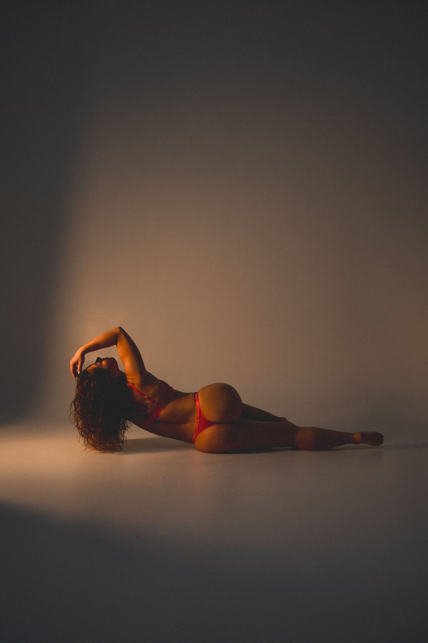 A woman in red bikini bottoms poses gracefully on the floor in dramatic warm studio lighting.
