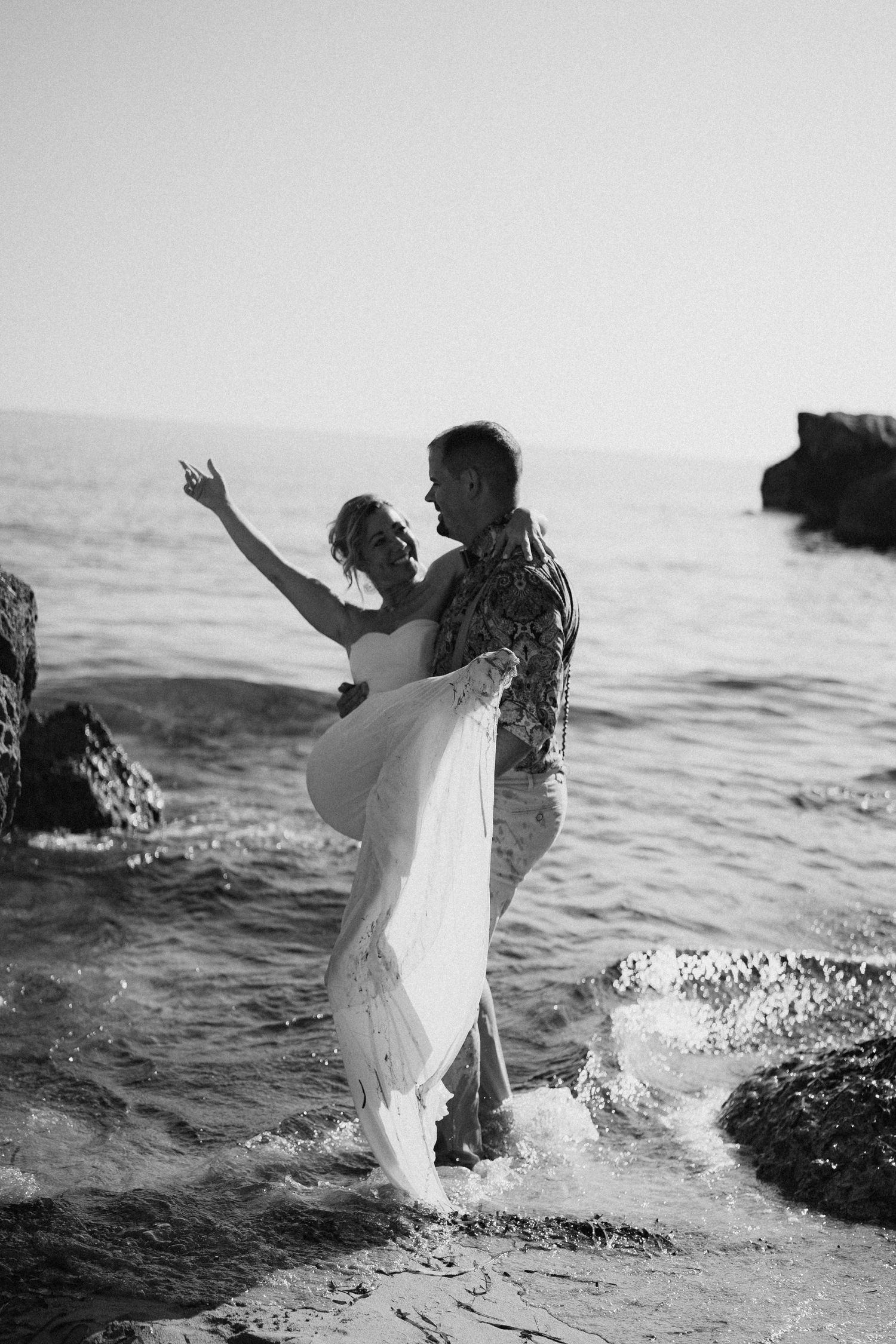 Black and white photo captures couple dancing playfully in ocean waves near rocky shore.