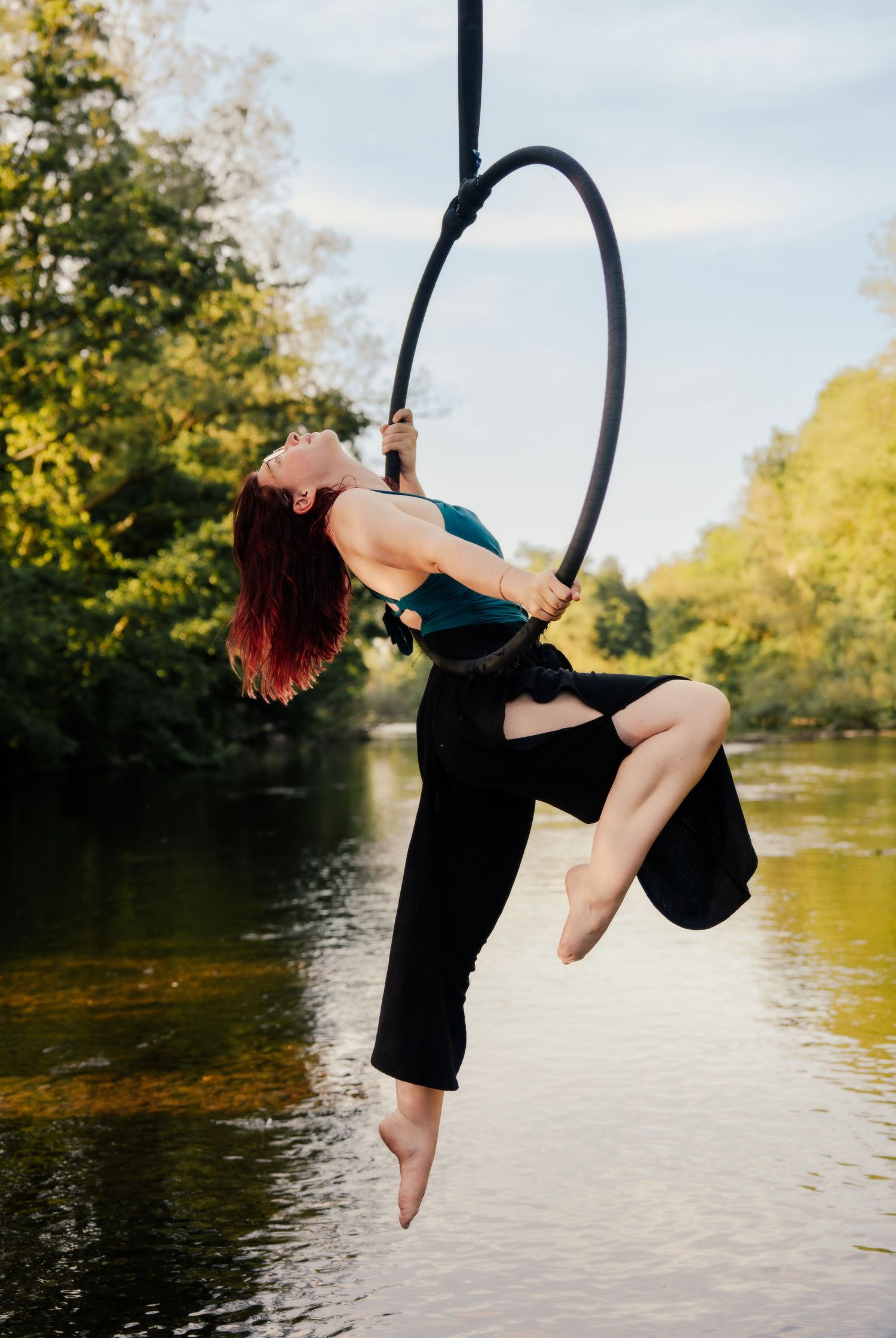 Woman performing aerial hoop lyra act outdoors over river, wearing teal top and black pants, surrounded by lush green trees.