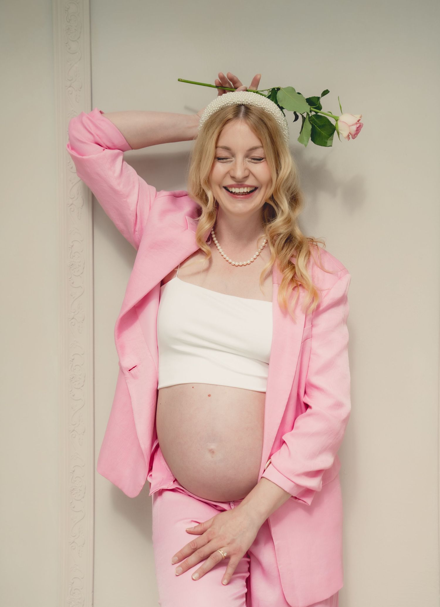 Smiling pregnant woman in pink blazer and pants with white crop top, pearl necklace, and floral headband.