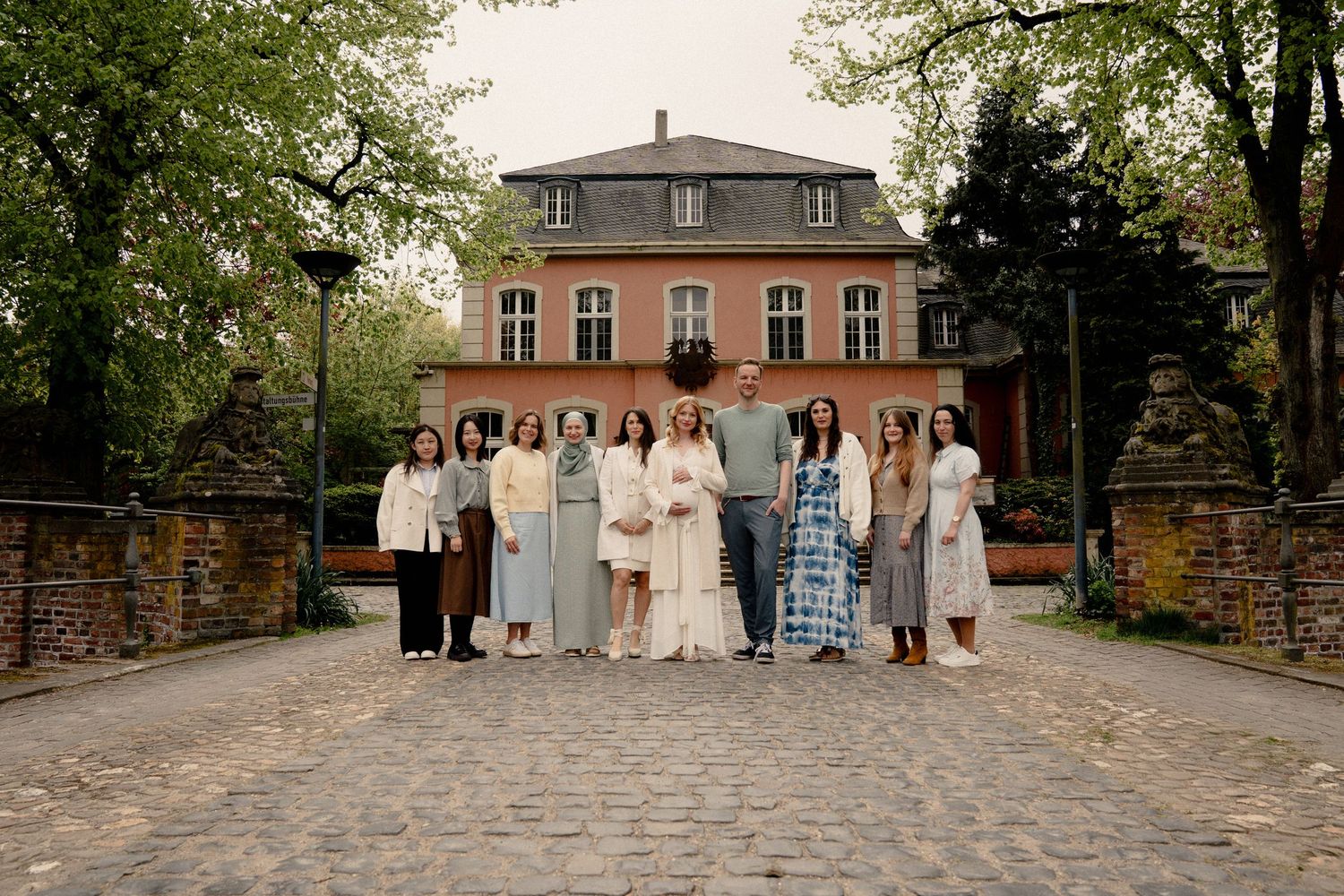 Group of women posing in front of a historic brick mansion surrounded by lush green trees on a cobblestone path.