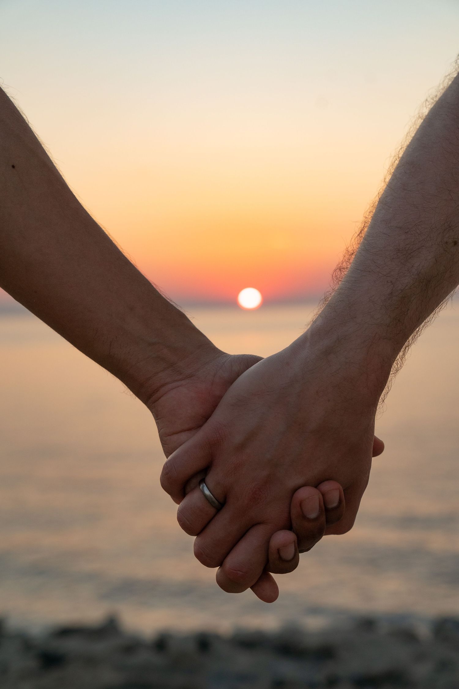 Couple holding hands with wedding rings at a romantic beach sunset, symbolizing love and togetherness.