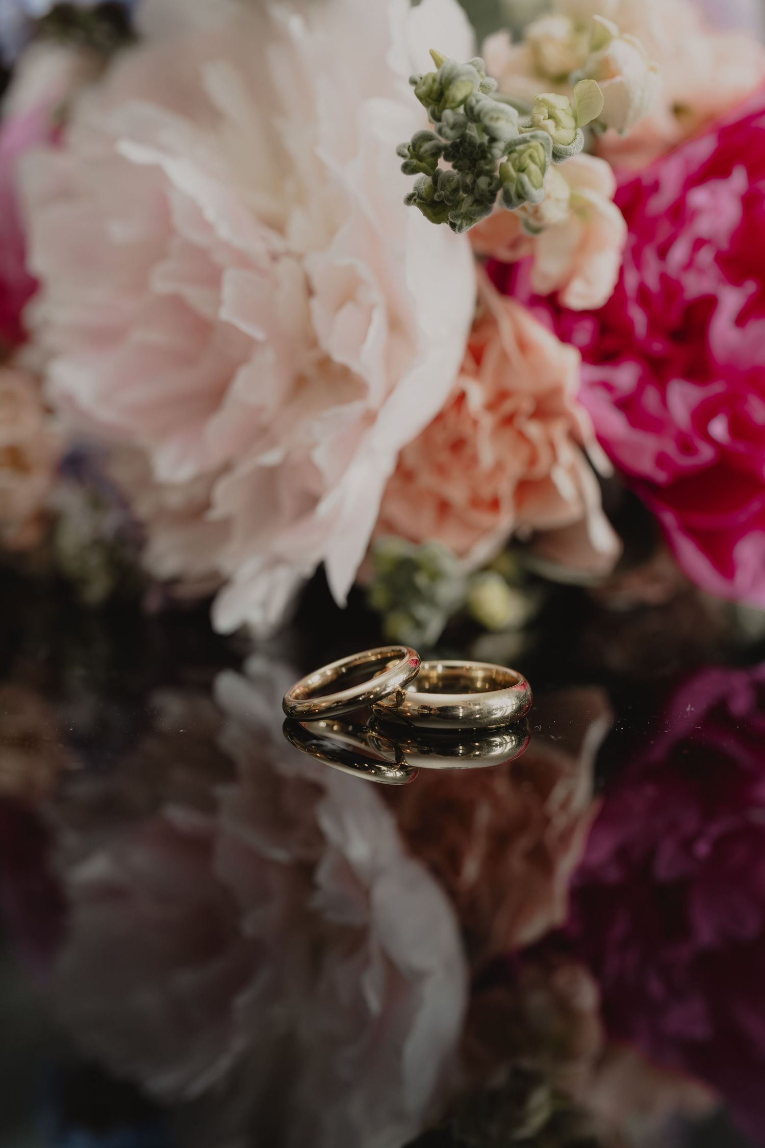 Two gold wedding bands rest on a reflective surface in front of a lush bouquet of pink and white peonies.