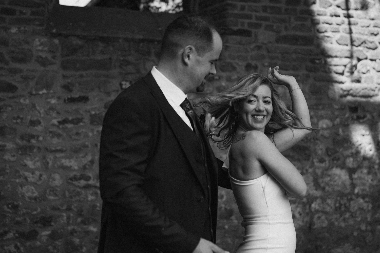 Black and white candid photo of joyful newlyweds sharing a lighthearted moment against brick wall.