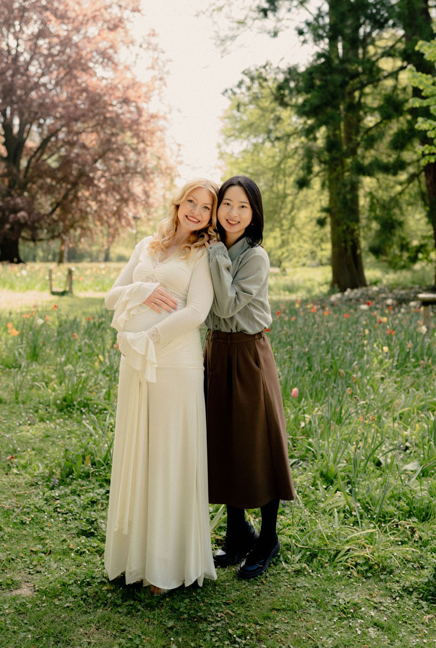 Pregnant woman in elegant white dress and friend smiling together in blooming spring park.