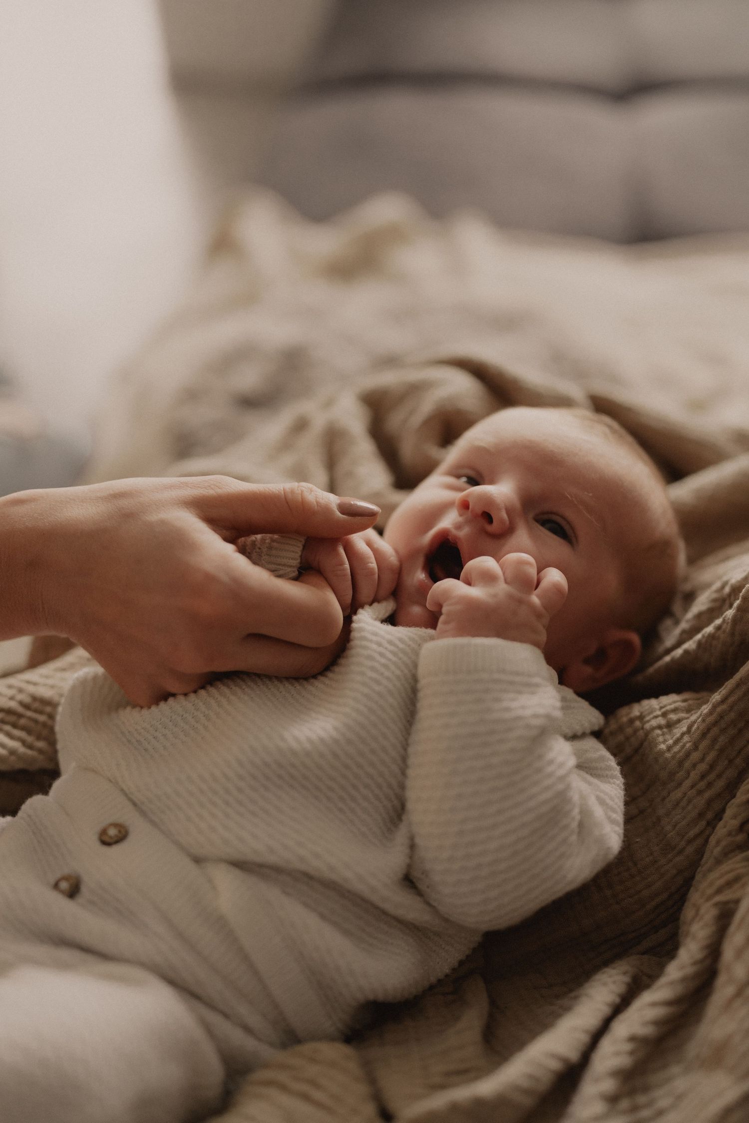Baby in white knit sweater lying on beige blanket with caring hands nearby.