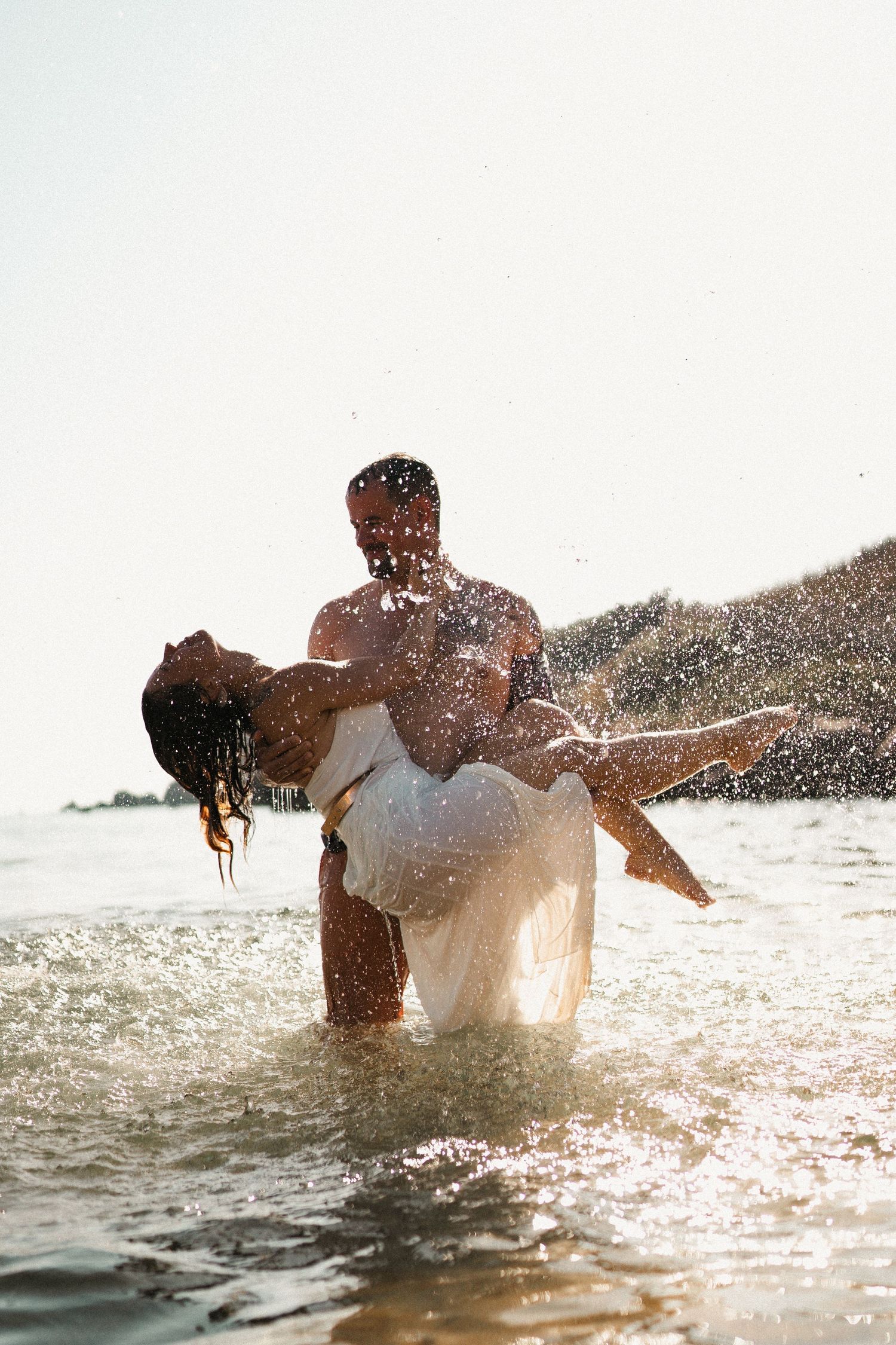 A couple plays in the sparkling ocean water during golden hour, creating dramatic splashes.