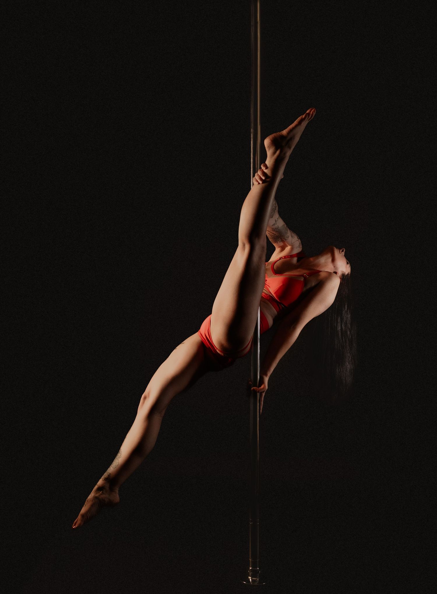 Flexible pole dancer in red outfit performs an aerial split pose on a vertical pole against a black background.
