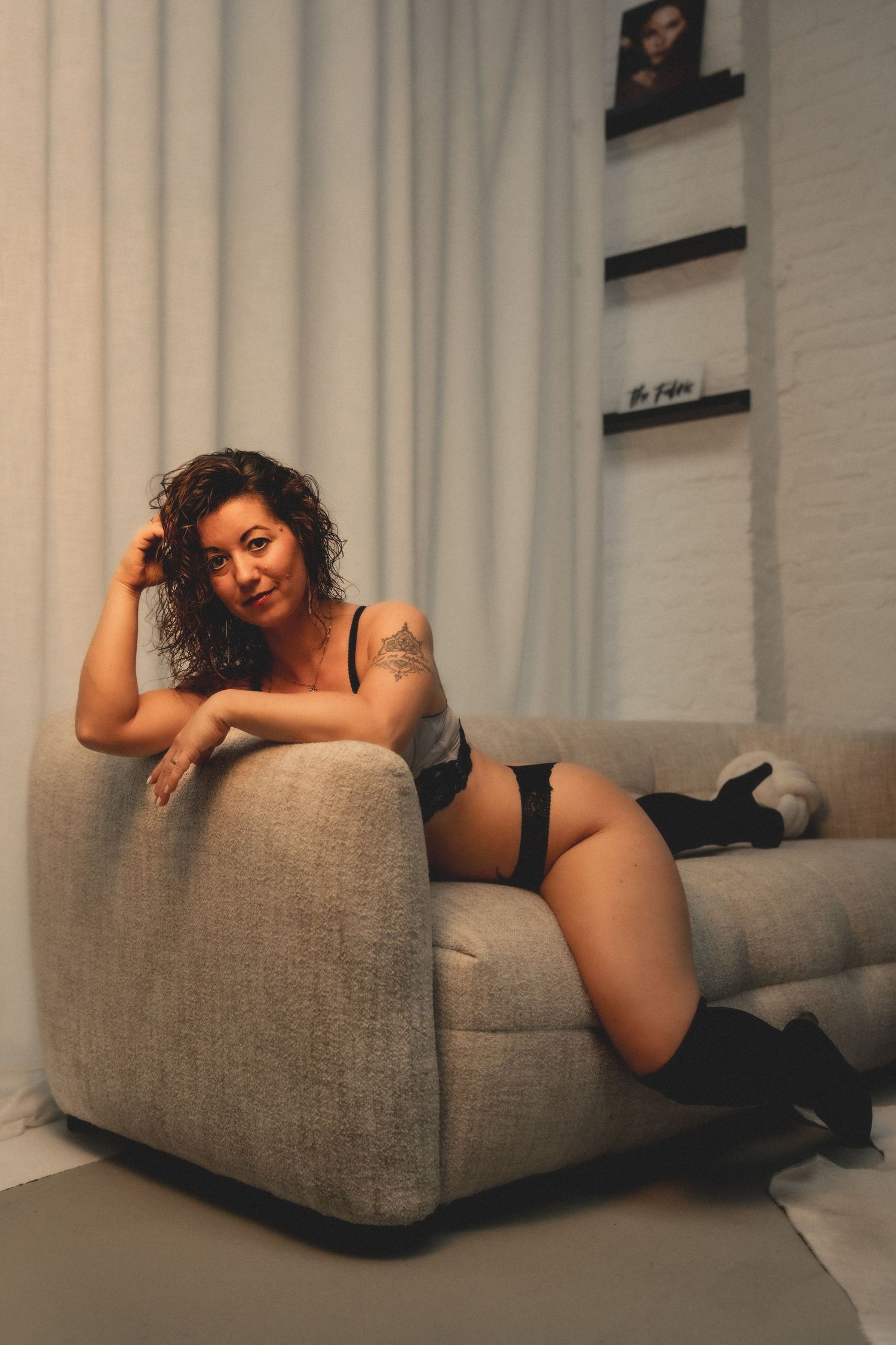 A curly-haired woman in black lingerie poses confidently beside a gray sofa in a warmly lit living room.
