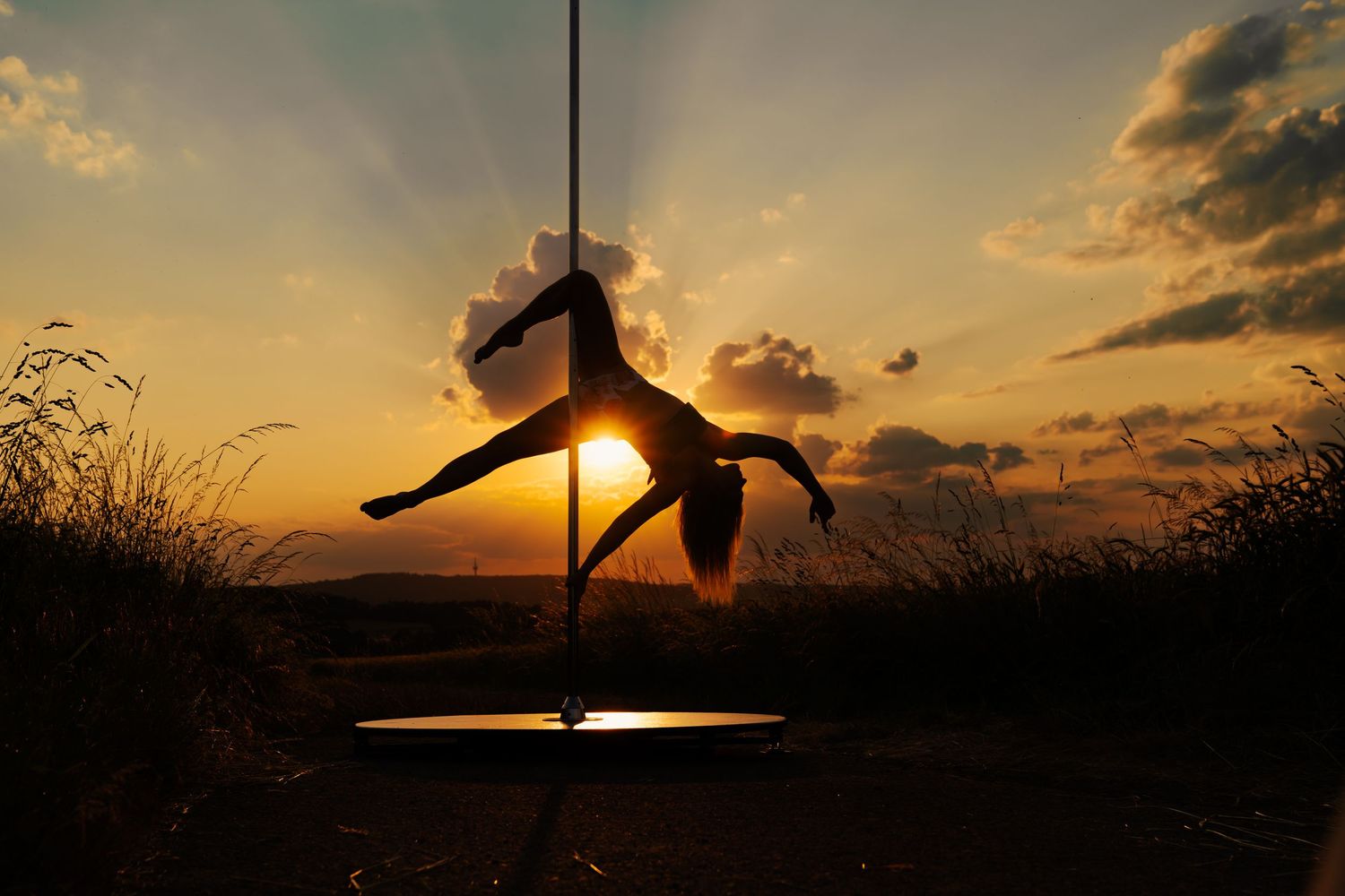 Silhouette of a pole dancer performing an acrobatic move outdoors against a stunning golden sunset sky.