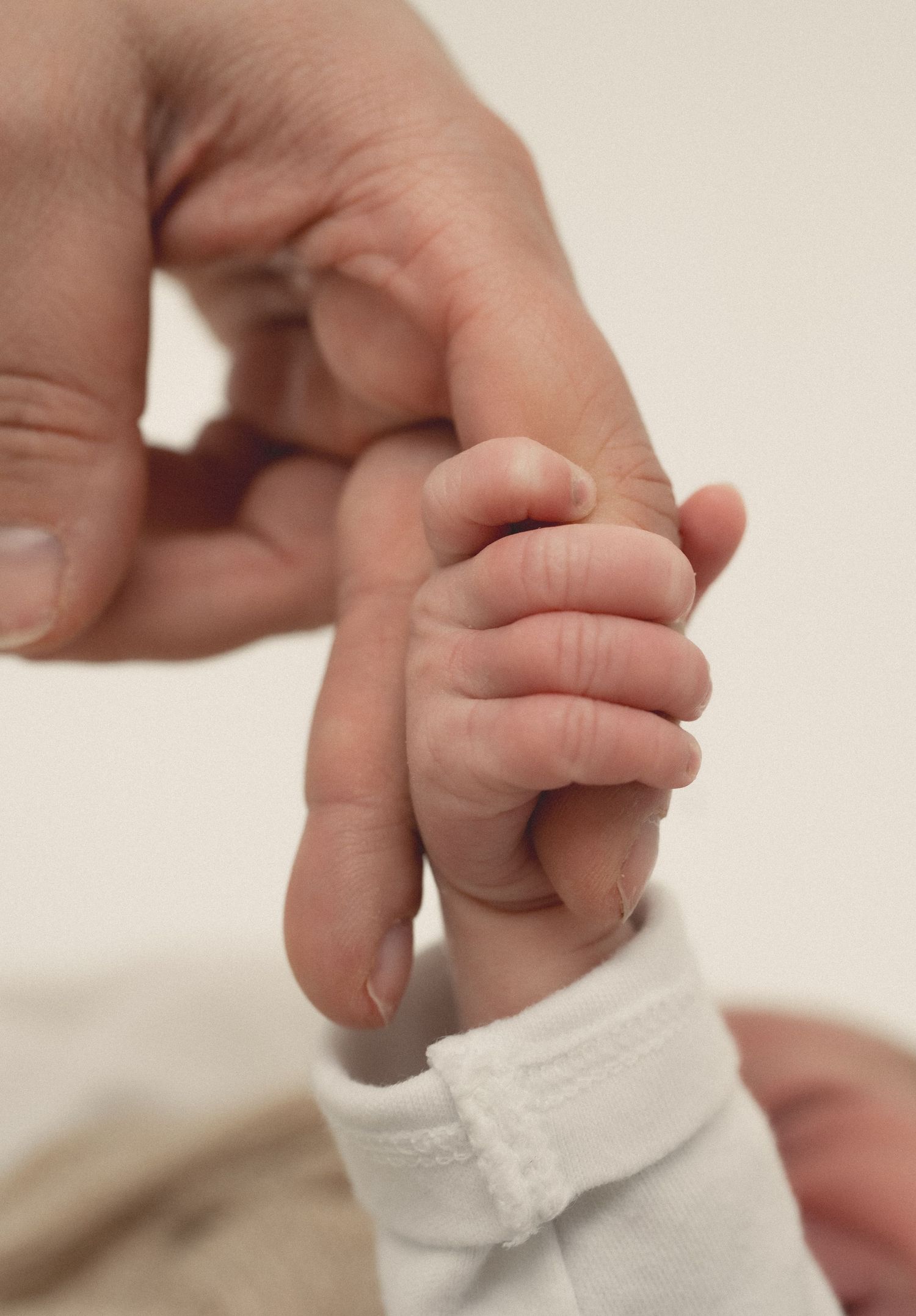 A newborn baby's tiny hand gripping an adult's finger, wearing a white onesie, symbolizing new life and parental bond.
