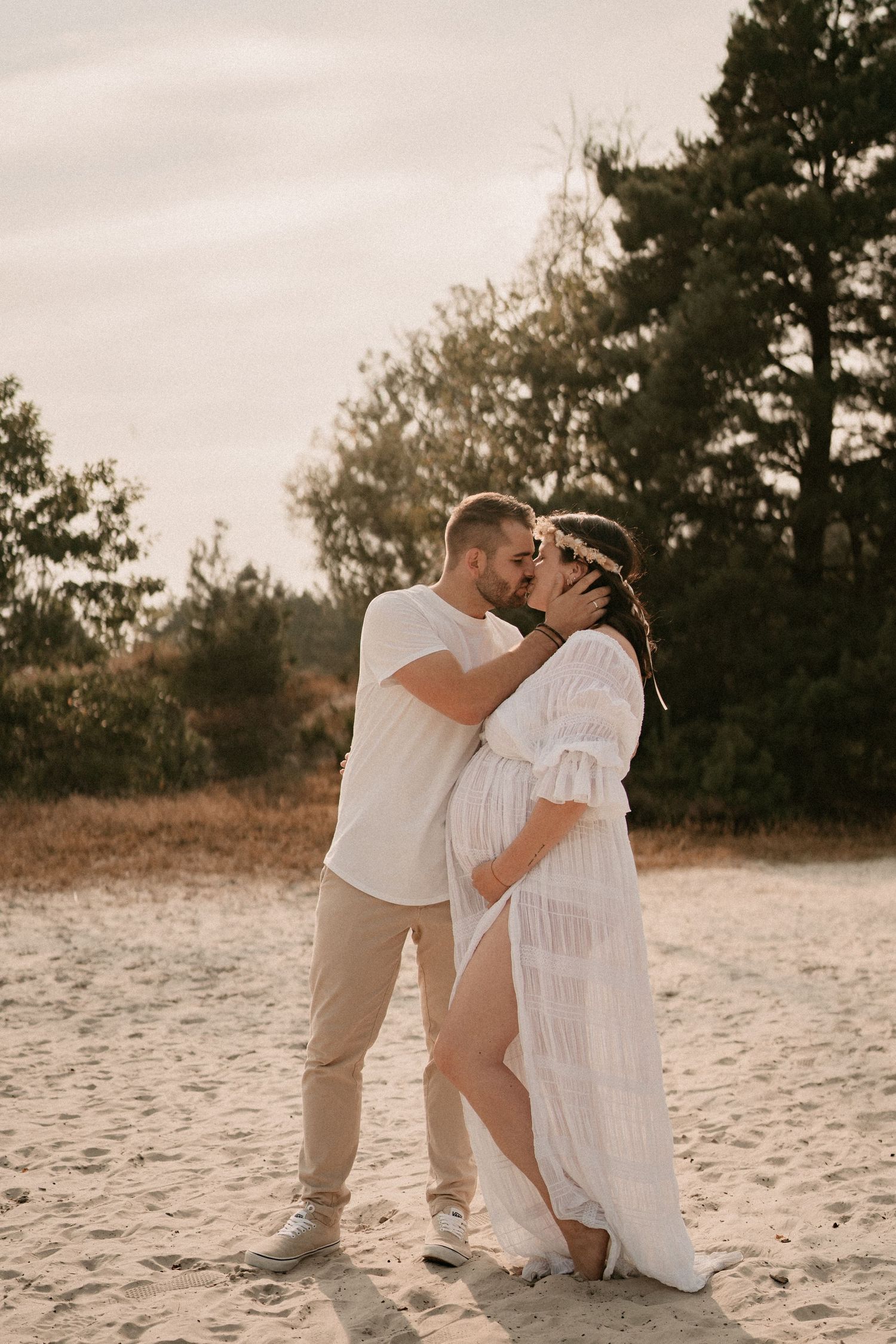 A romantic beach photoshoot with a couple in white outfits embracing on sandy dunes at sunset.
