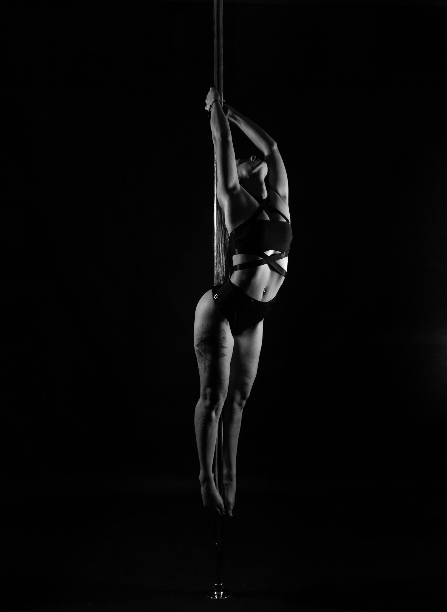 Black and white photo of a pole dancer performing an artistic vertical hold on a pole against a dark background.