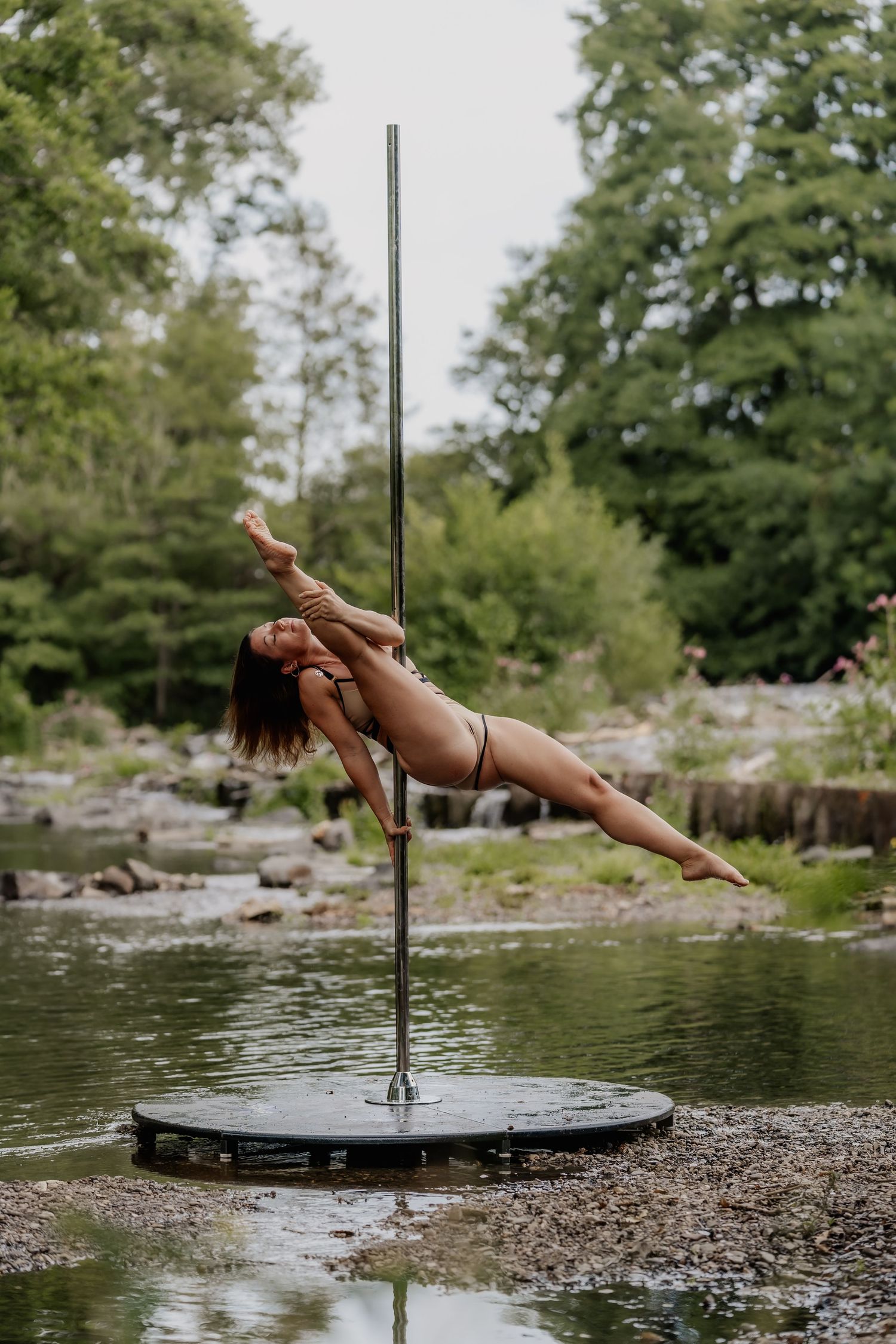 Woman performing aerial pole dance move outdoors near rocky stream surrounded by lush green trees.