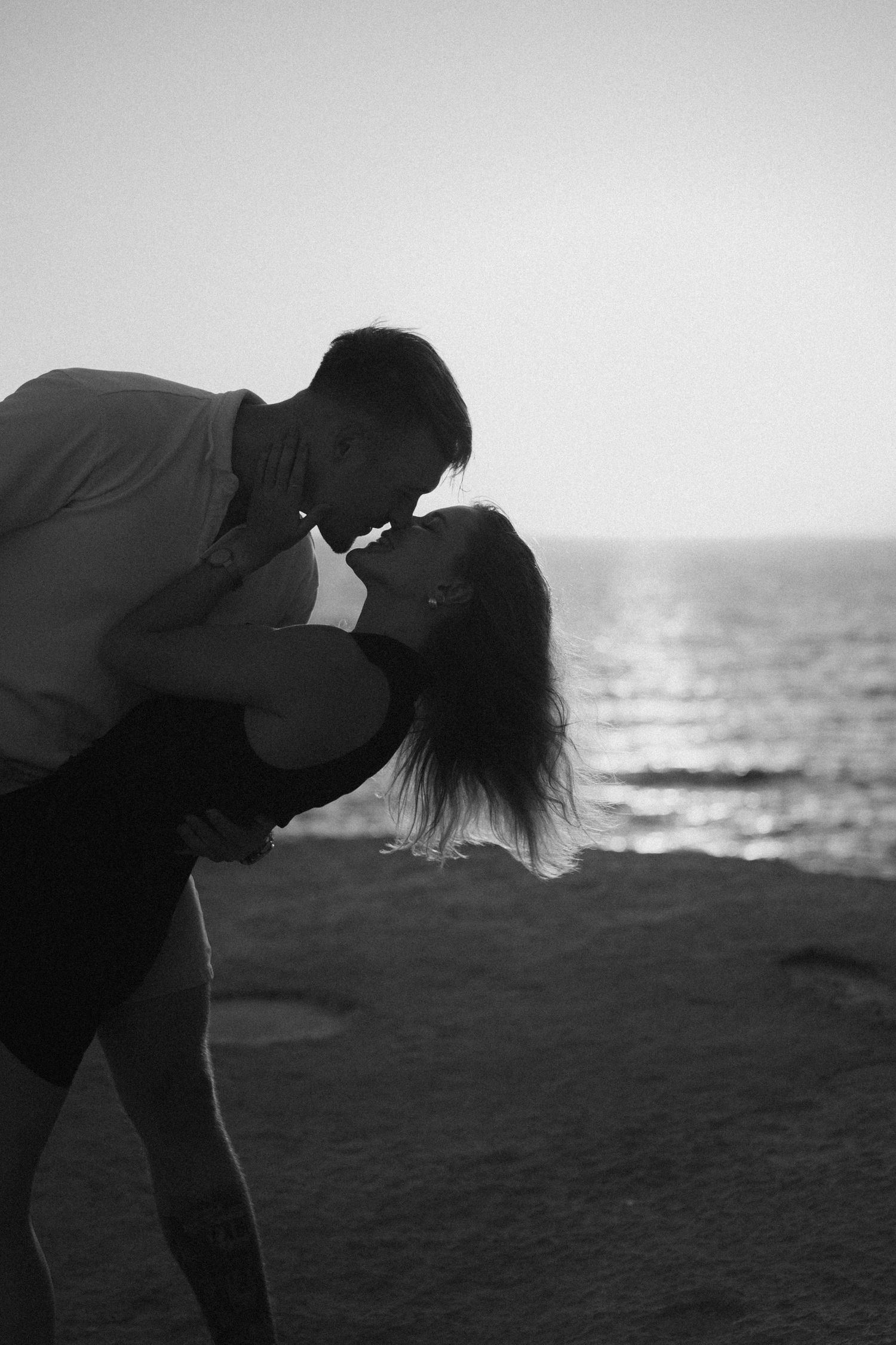 Black and white photo of a romantic couple kissing on a beach at sunset, silhouetted against the ocean.