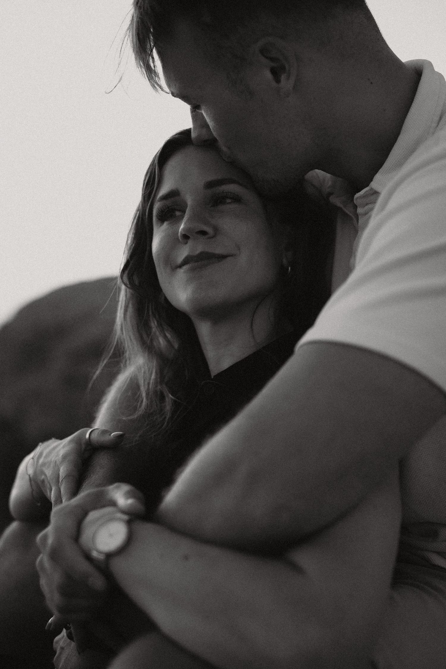 Black and white photo of a couple embracing, man kissing woman's forehead while she smiles softly.