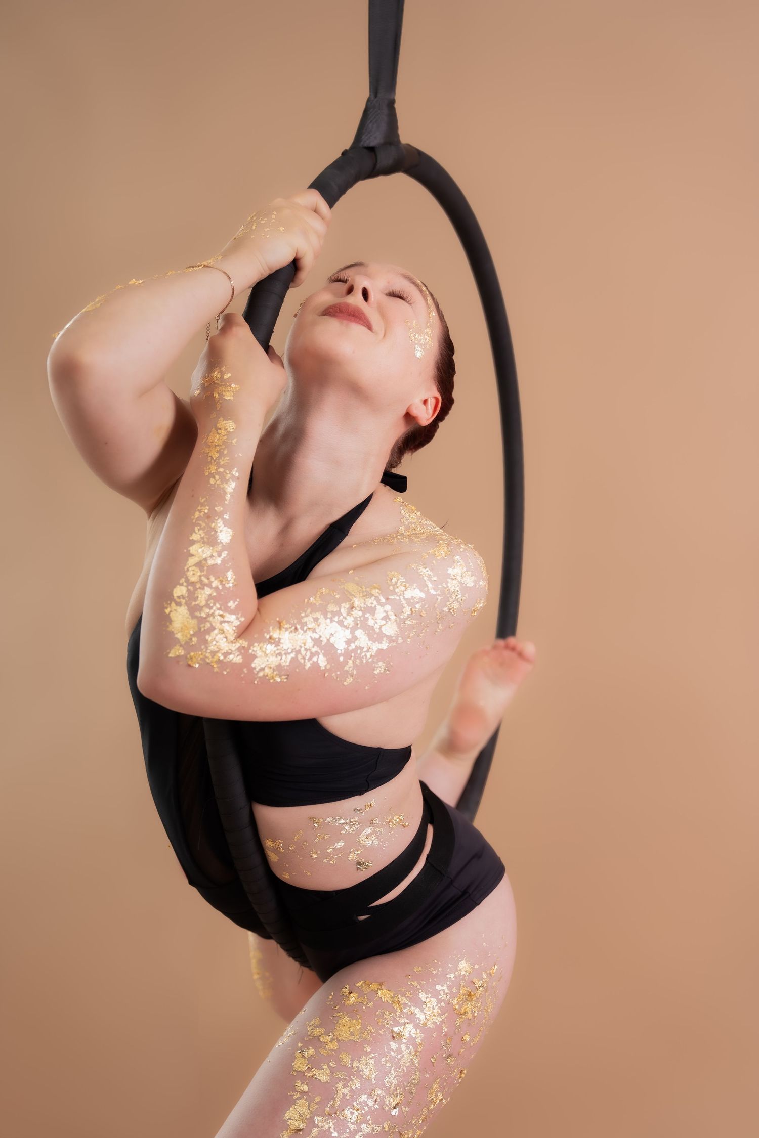 Woman with gold glitter performs aerial hoop routine in black activewear against beige background.