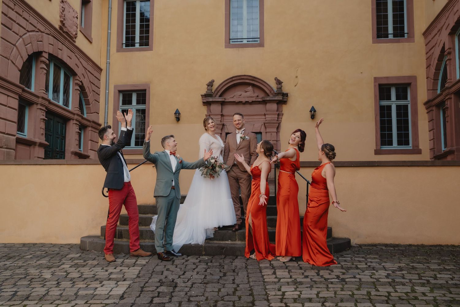 Wedding party in red bridesmaid dresses poses in front of a yellow stone building.