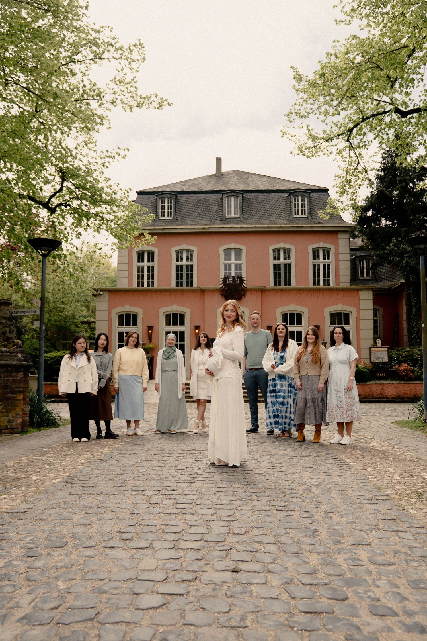 Pregnant woman in white lace gown poses with group of guests in front of a pink European manor on cobblestone courtyard.