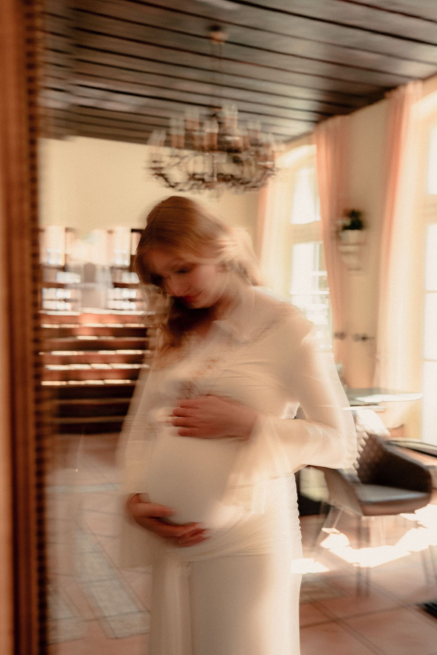 Dreamy blurred reflection of a pregnant woman in white dress cradling her belly in a sunlit vintage interior.