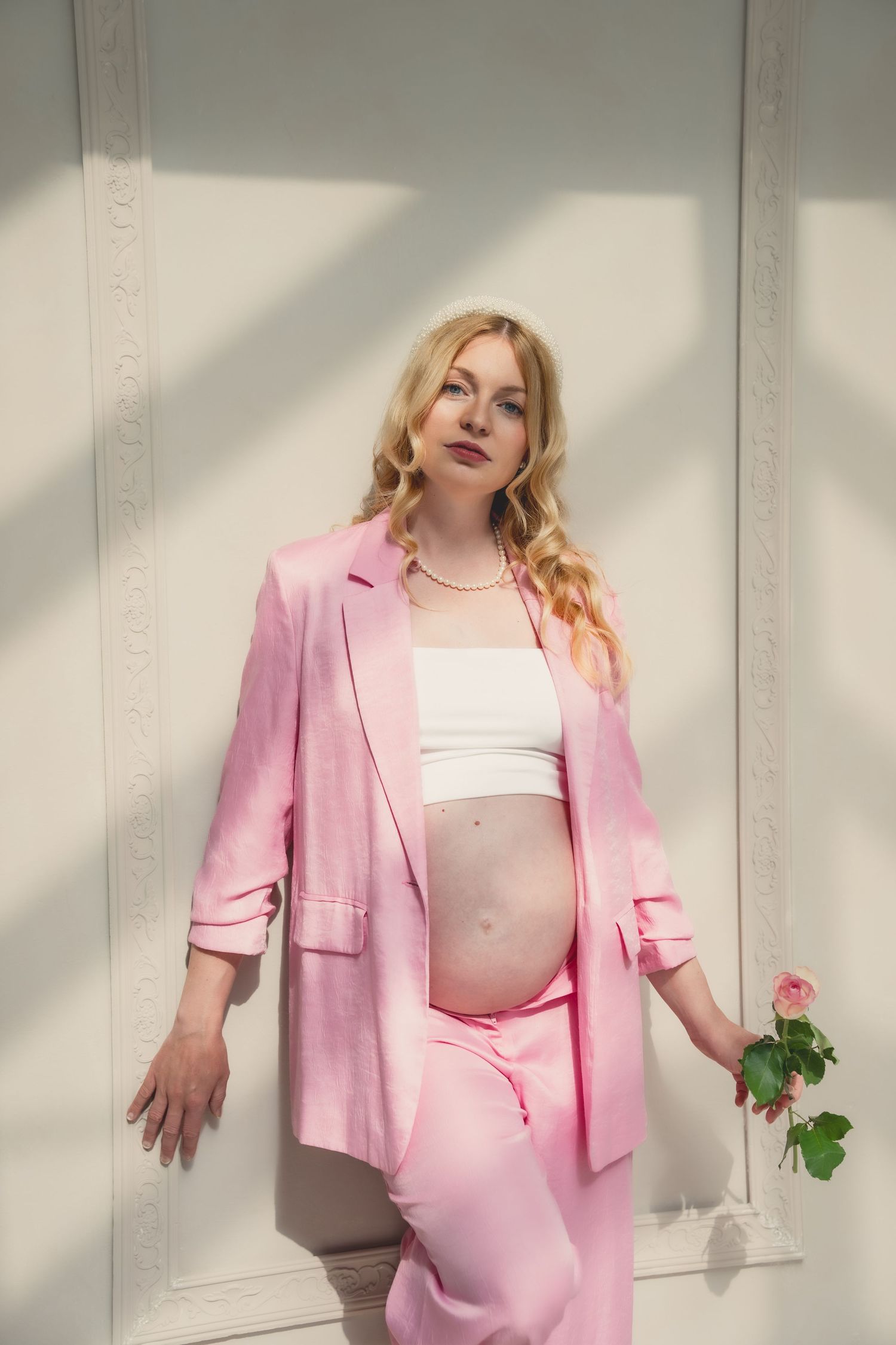 Pregnant woman in pink blazer and pants holding a rose, posing against a white wall in natural light.