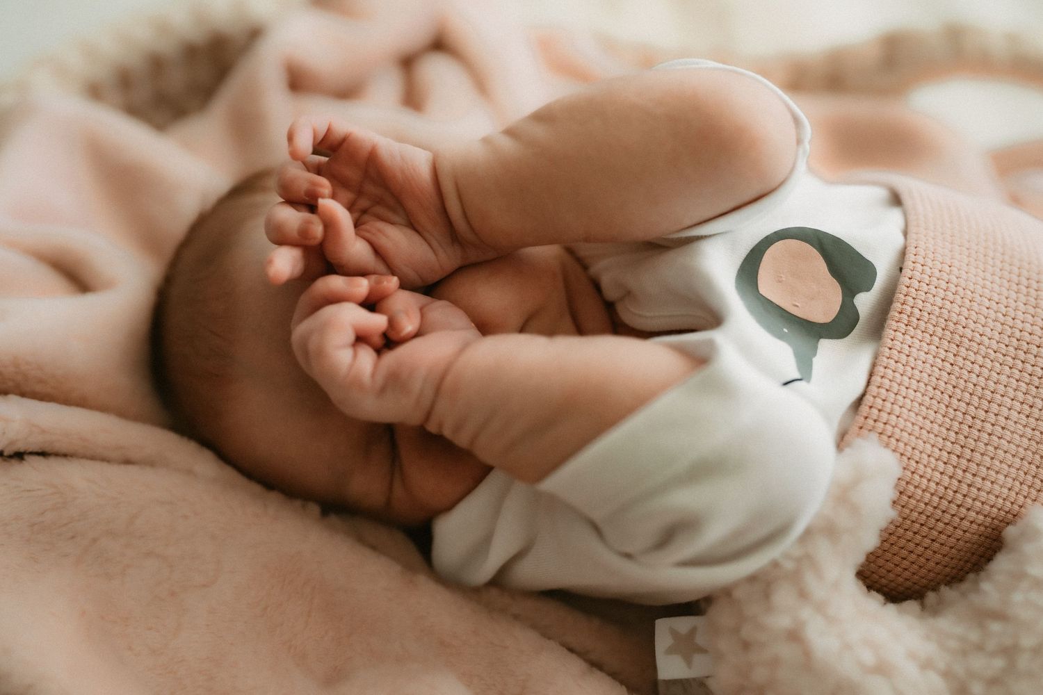 A newborn baby's tiny hand grasps a finger while resting on soft white bedding.
