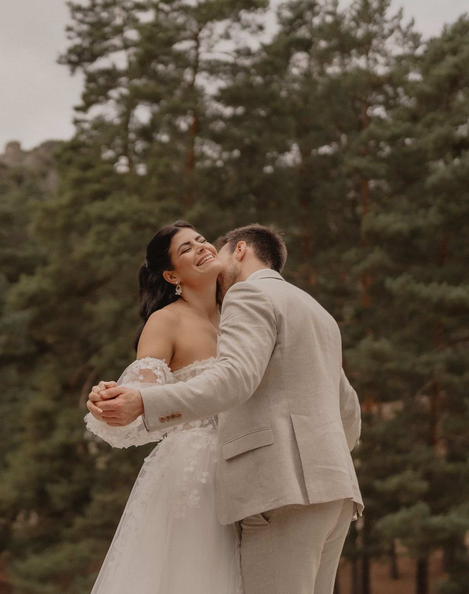 A couple shares a tender moment in a pine forest wearing wedding attire.