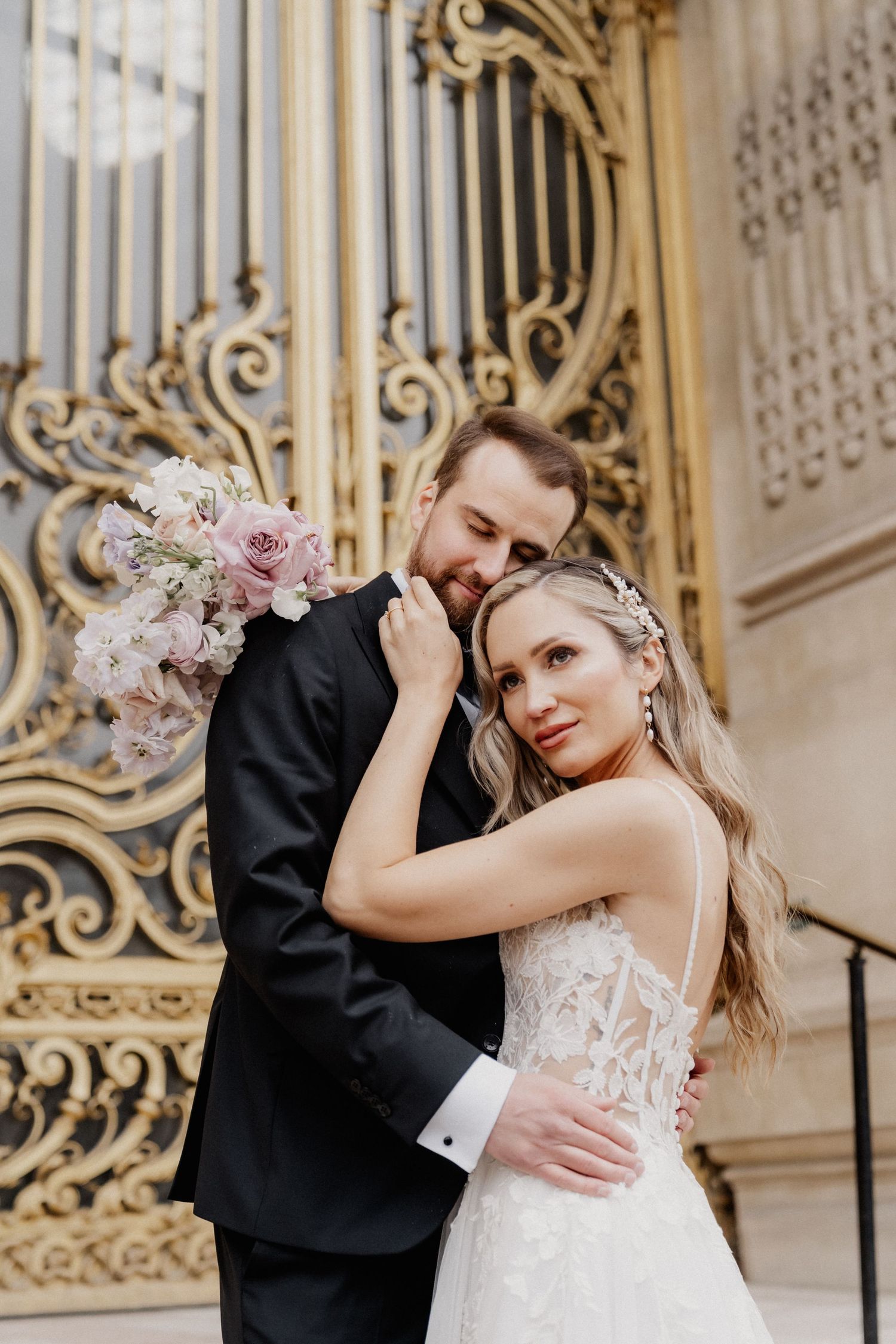 A romantic wedding couple embraces in front of ornate golden gates with delicate floral details and architectural elements.